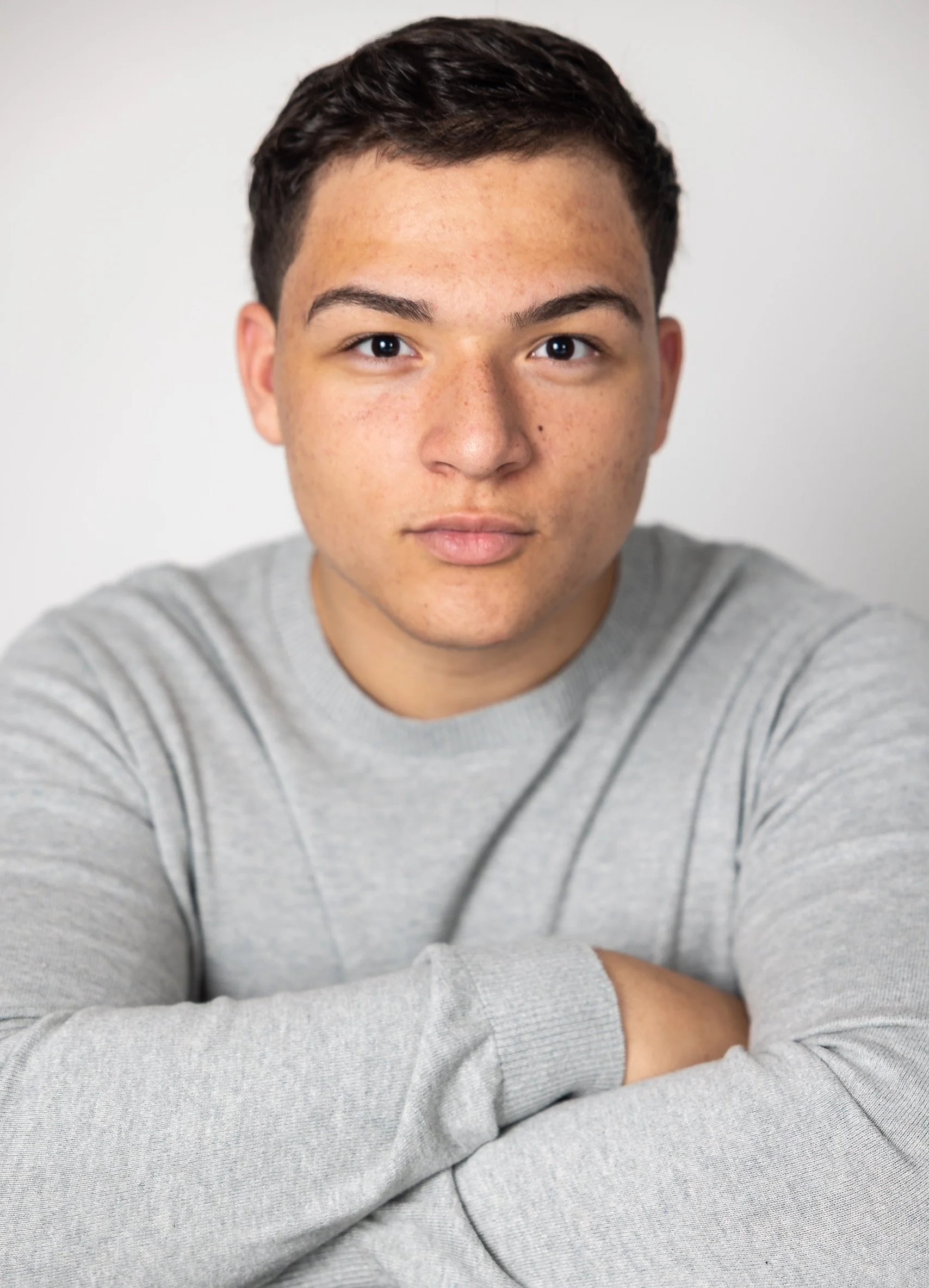 Close-up portrait of a young man with short dark hair, brown eyes, and a light complexion, wearing a gray sweater, with arms crossed in front of a neutral background.