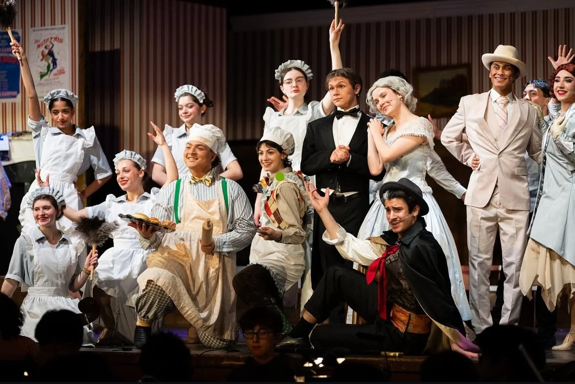 Cast of a theatrical performance dressed in costumes on stage, including chefs, a waiter, and elegant dresses, in a curtain-lined theater setting.