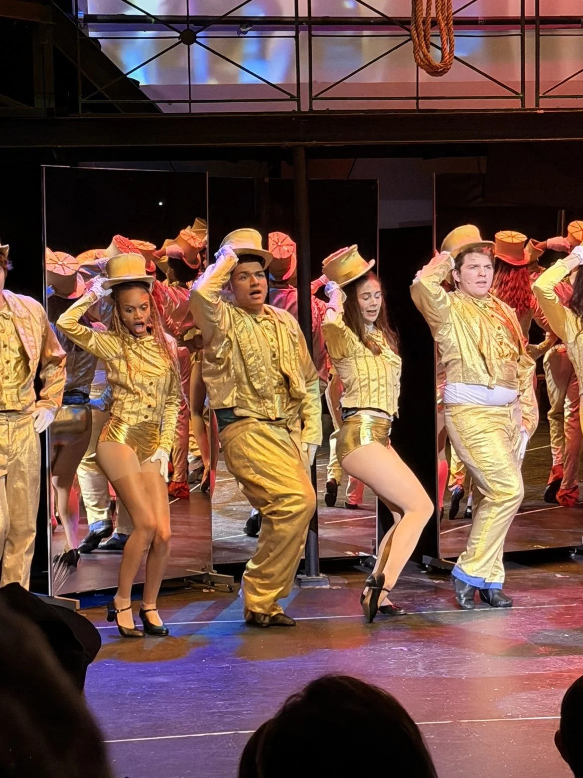 Male and female dancers performing on stage in gold costumes, with some wearing hats and high heels, surrounded by mirrors, during a stage show.