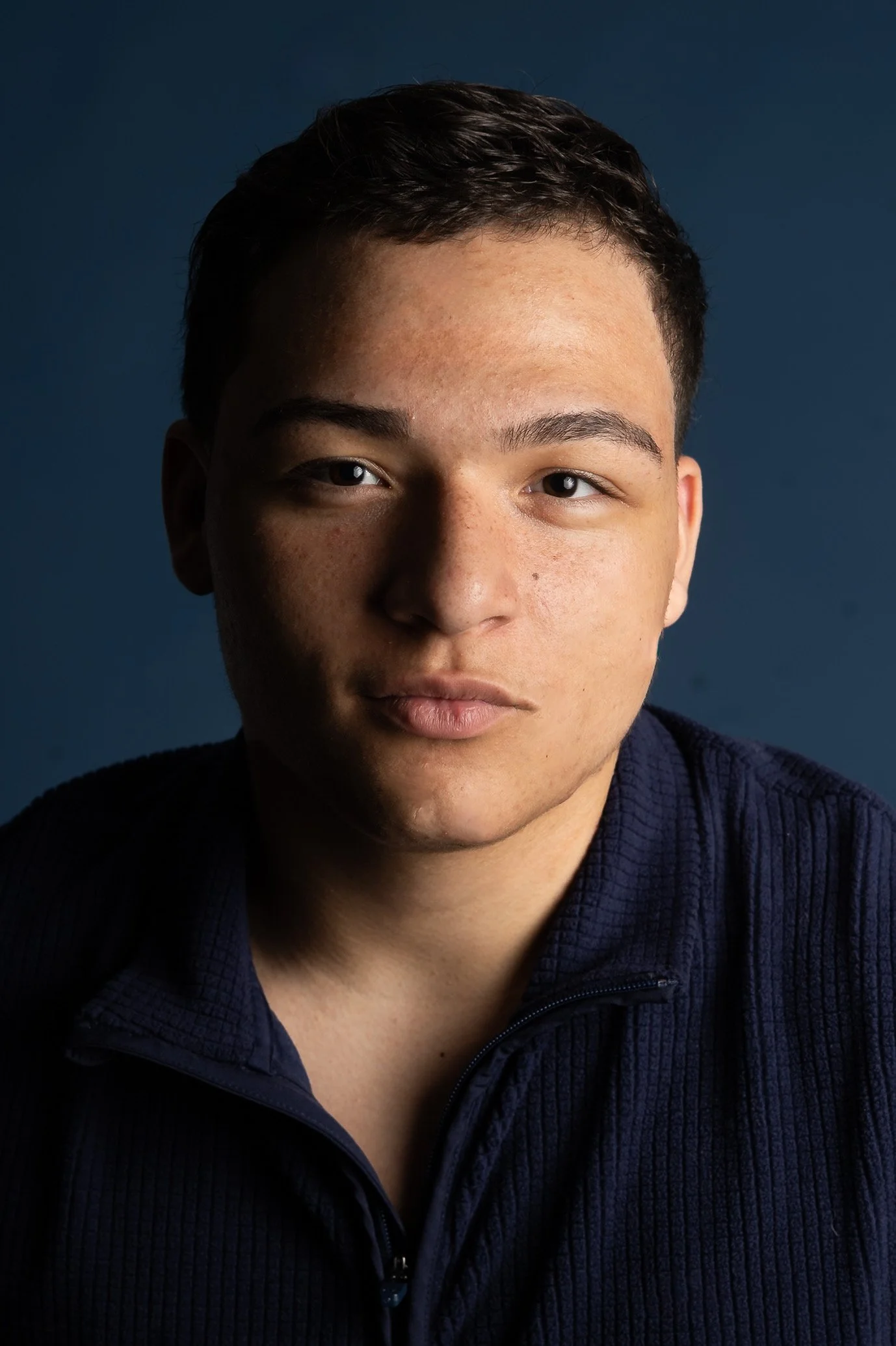 Close-up portrait of a young man with short dark hair, brown eyes, and a neutral expression, wearing a dark navy zip-up sweater against a dark background.