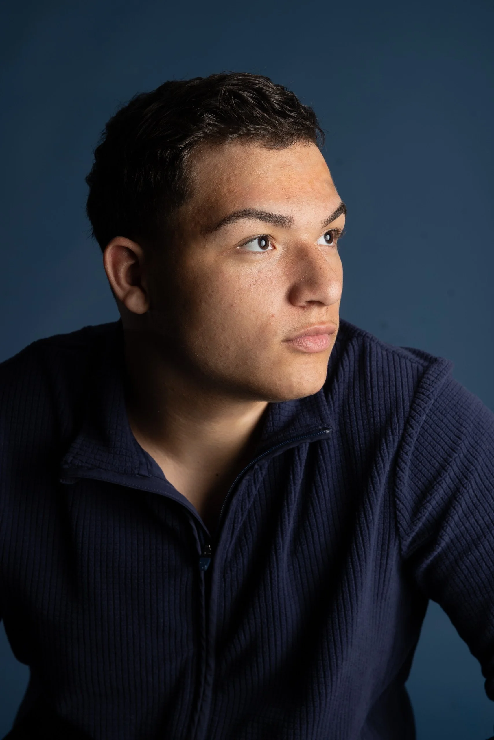 A young man with short dark hair and light skin, looking slightly to the right, wearing a navy blue textured zip-up jacket against a dark blue background.