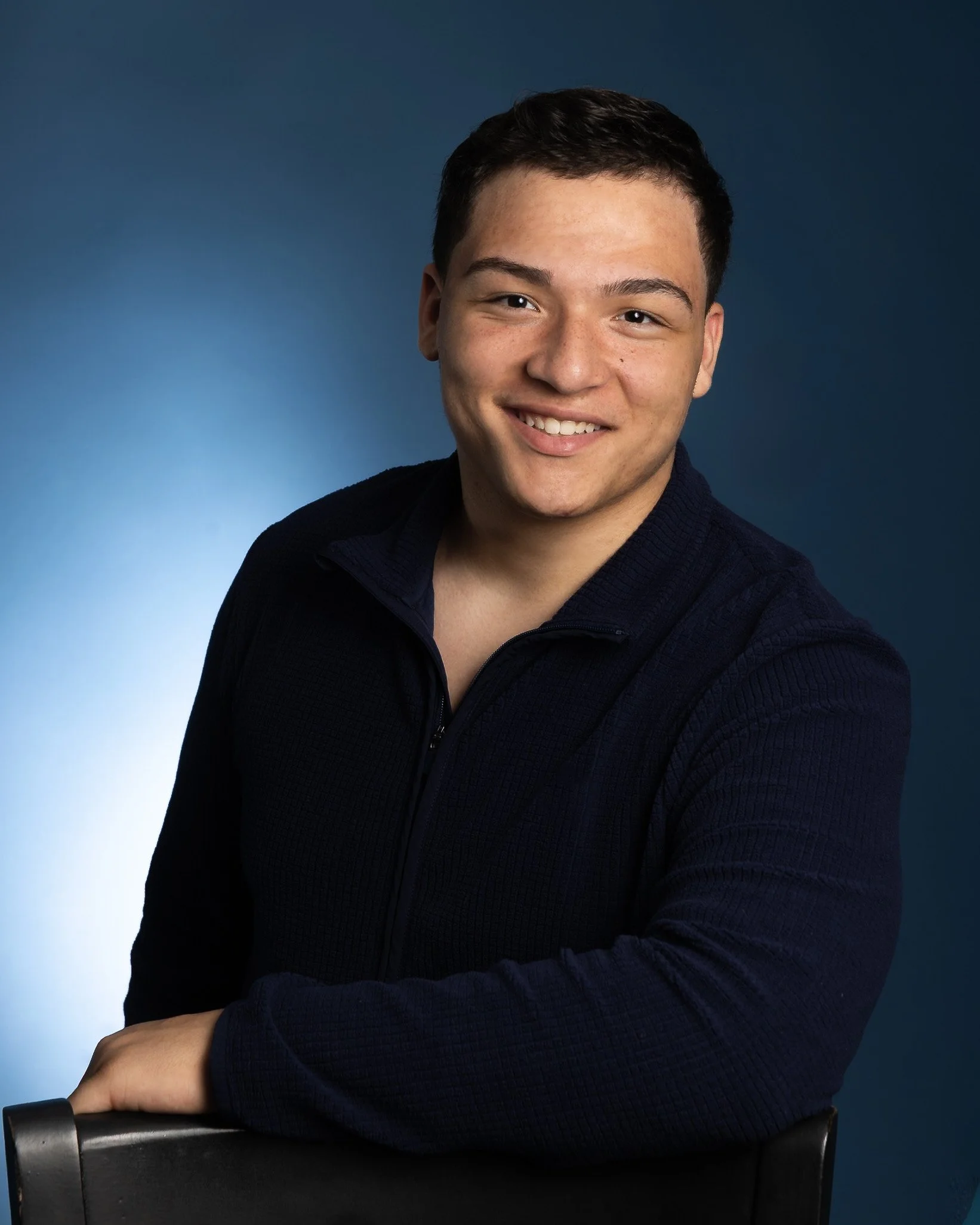 A young man with short dark hair, wearing a dark sweater, sitting against a gradient blue background, smiling at the camera with his arms crossed.