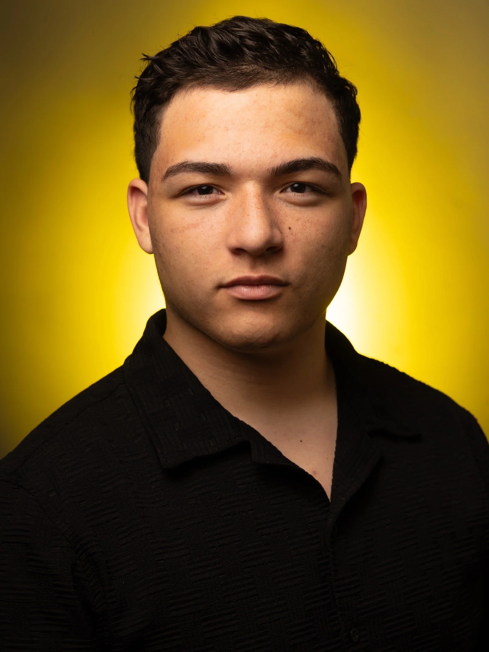 Close-up portrait of a young man with short dark hair, wearing a black textured shirt, against a yellow background.
