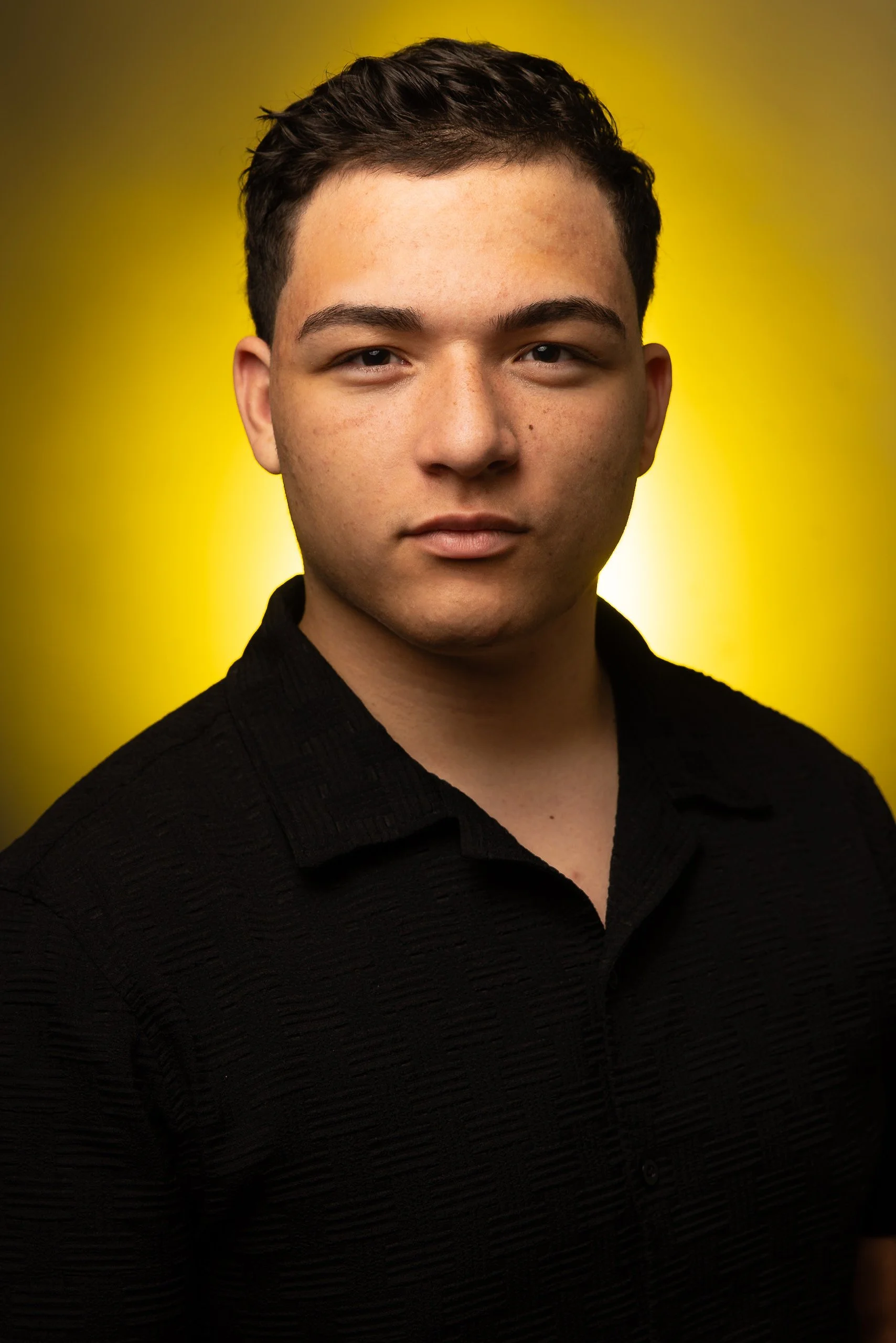 A young man with short dark hair and a light complexion, wearing a black textured shirt, standing against a yellow background.