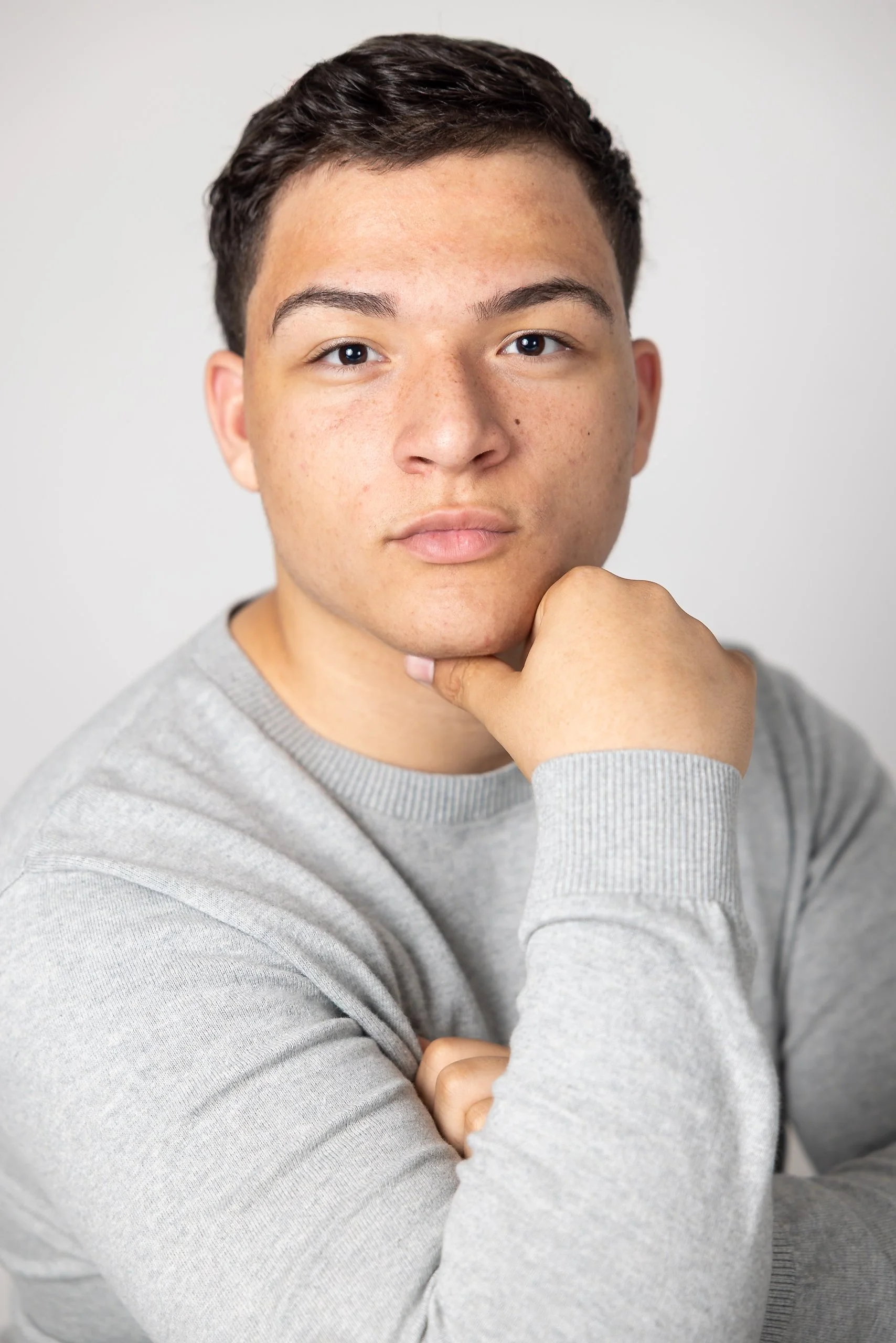 A young man with short dark hair and light brown skin, wearing a grey sweatshirt, resting his chin on his hand with a neutral expression, against a plain light grey background.