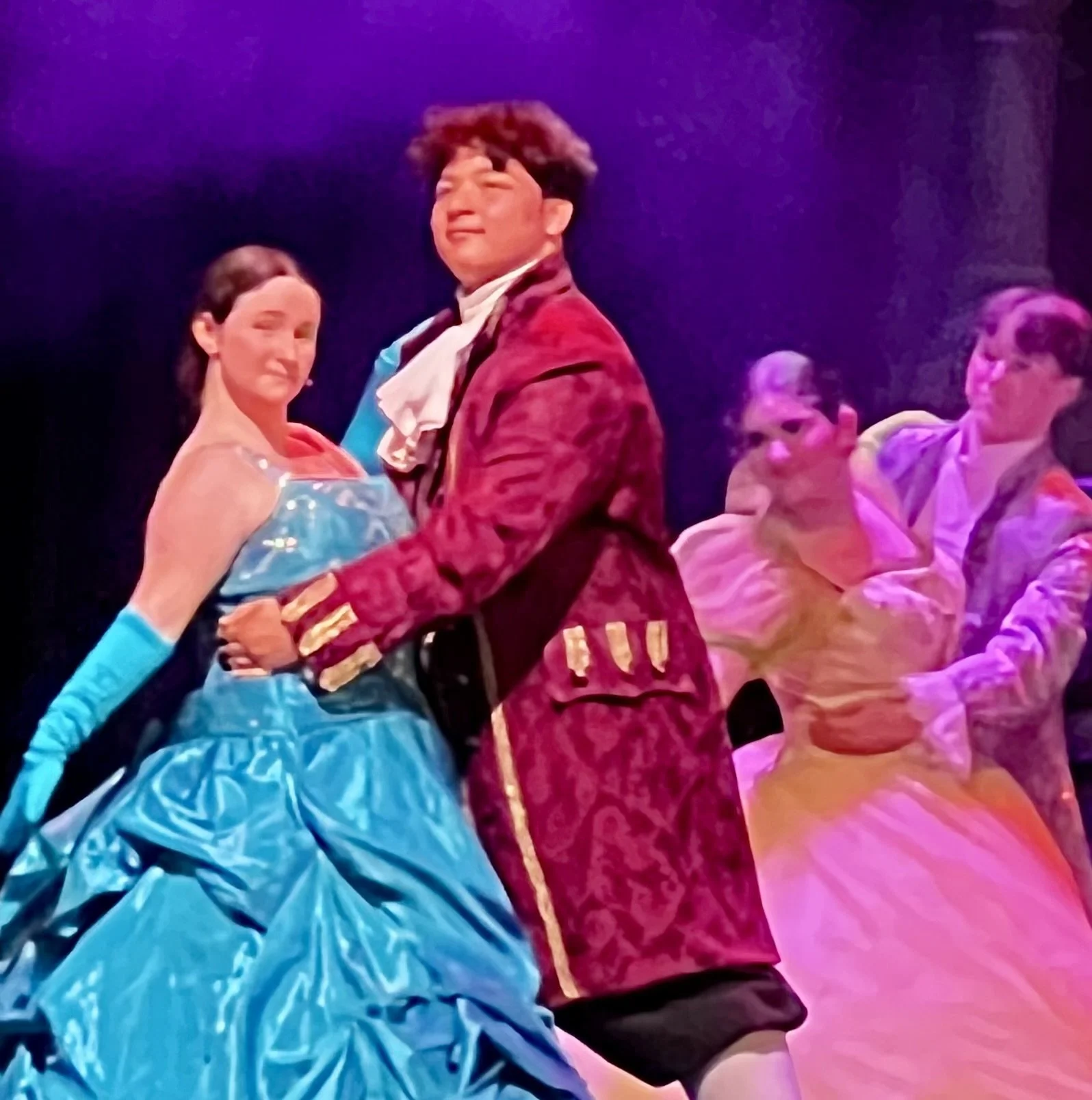 The image depicts four performers on a stage, dressed in colorful costumes with a purple background and vibrant lighting. One performer at the center is dressed in a red and gold regal-looking costume, while another to her left is in a shiny blue dre