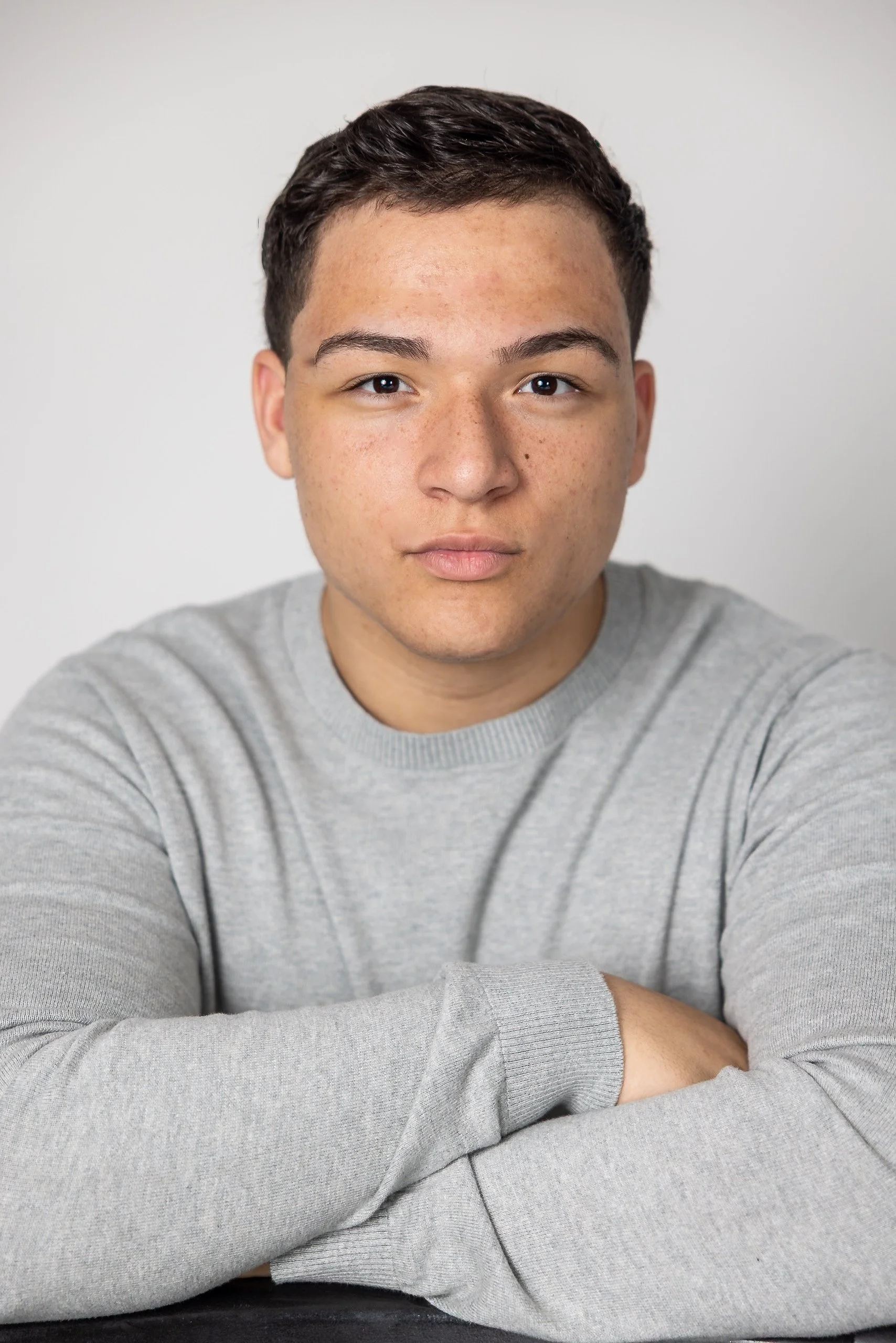 Close-up of a young man with dark hair and brown eyes wearing a gray sweater, resting his arms on a dark surface against a plain white background.
