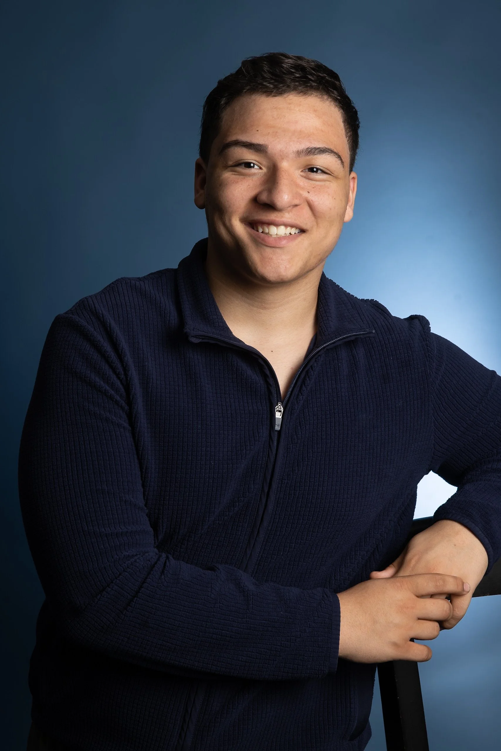 A young man with short dark hair smiling, wearing a navy blue zip-up sweater, and sitting with one arm resting on a chair against a blue gradient background.