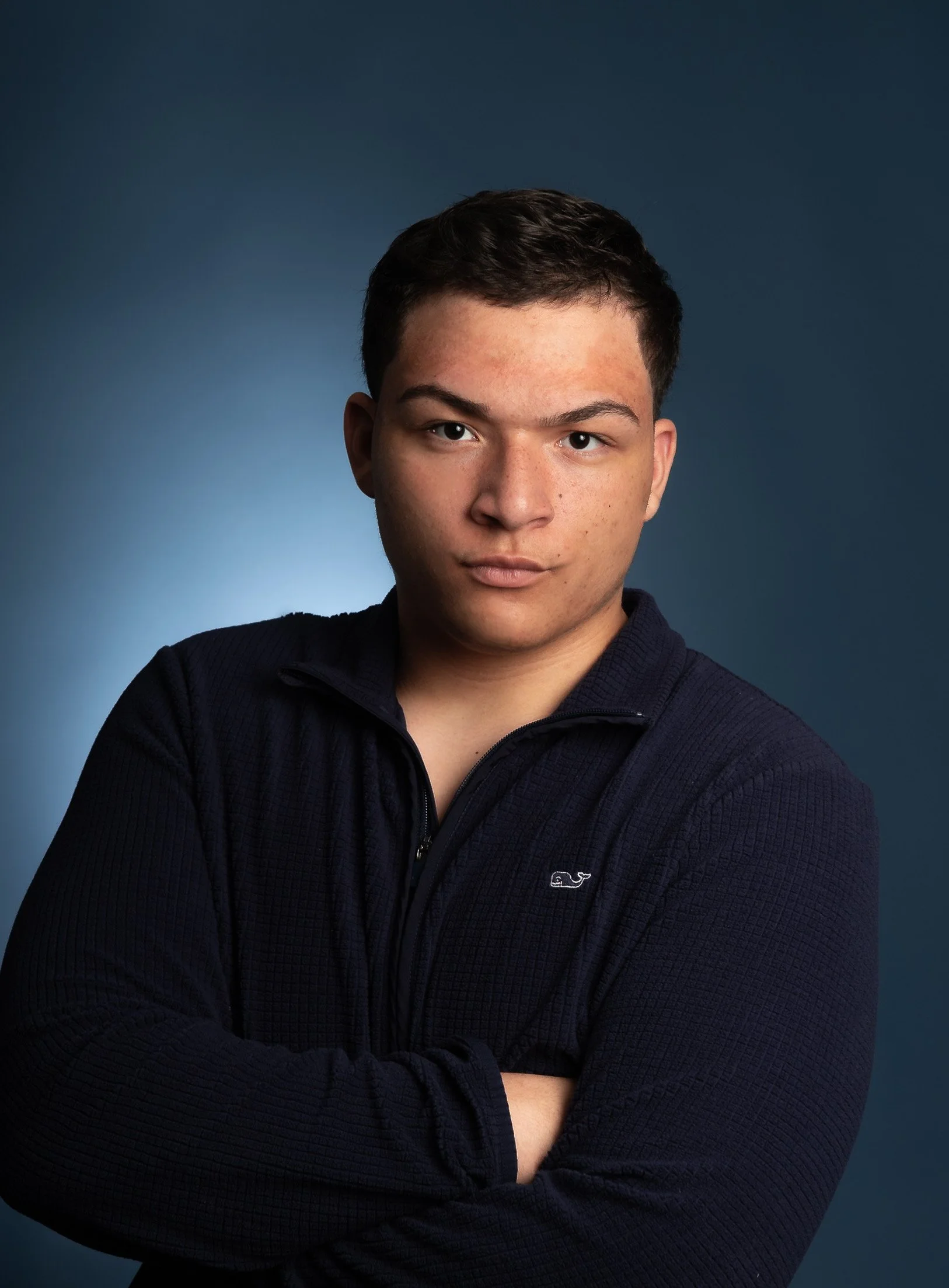 A young man with short dark hair, wearing a dark zip-up sweater with a small logo on the chest, posing with crossed arms against a dark blue background.