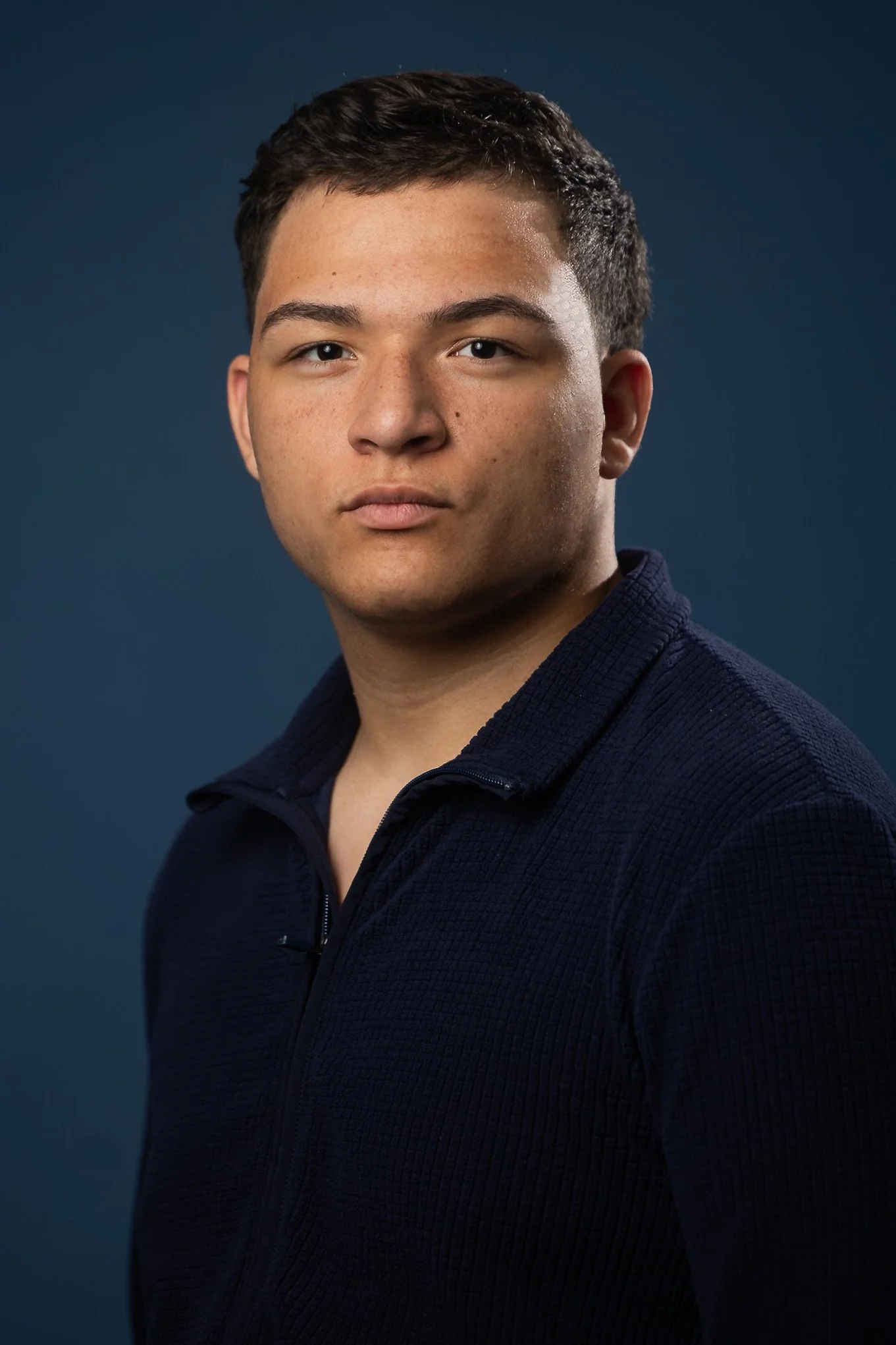 Close-up portrait of a young man with short dark hair, fair skin, and brown eyes, wearing a dark zip-up jacket against a dark blue background.