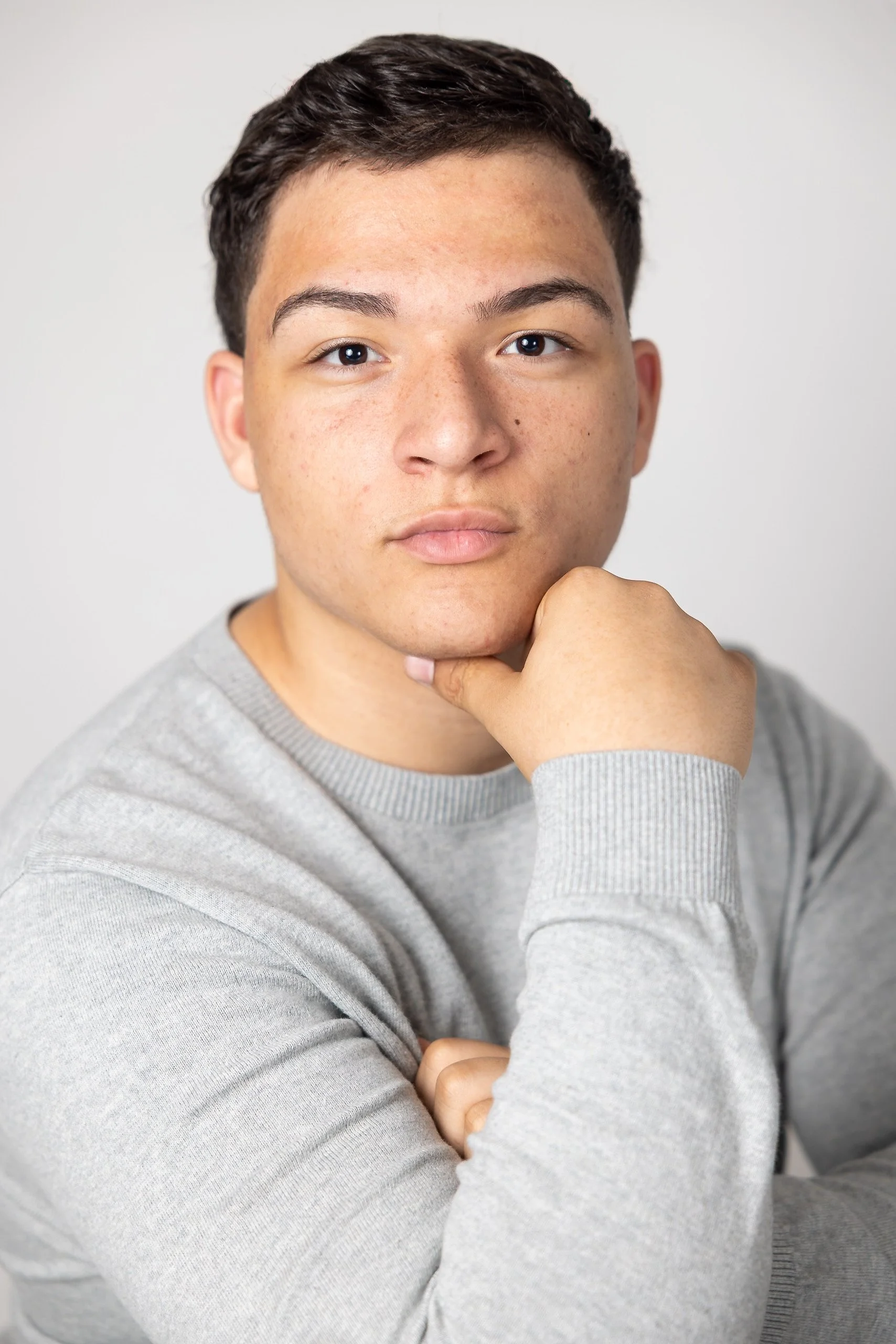 A young man with short dark hair and light brown skin, wearing a light gray sweatshirt, rests his chin on his hand while looking at the camera against a plain white background.