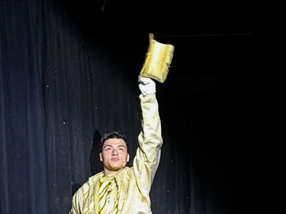 A man in a shiny yellow jacket and white gloves holding a yellow trophy above his head in front of a black curtain.