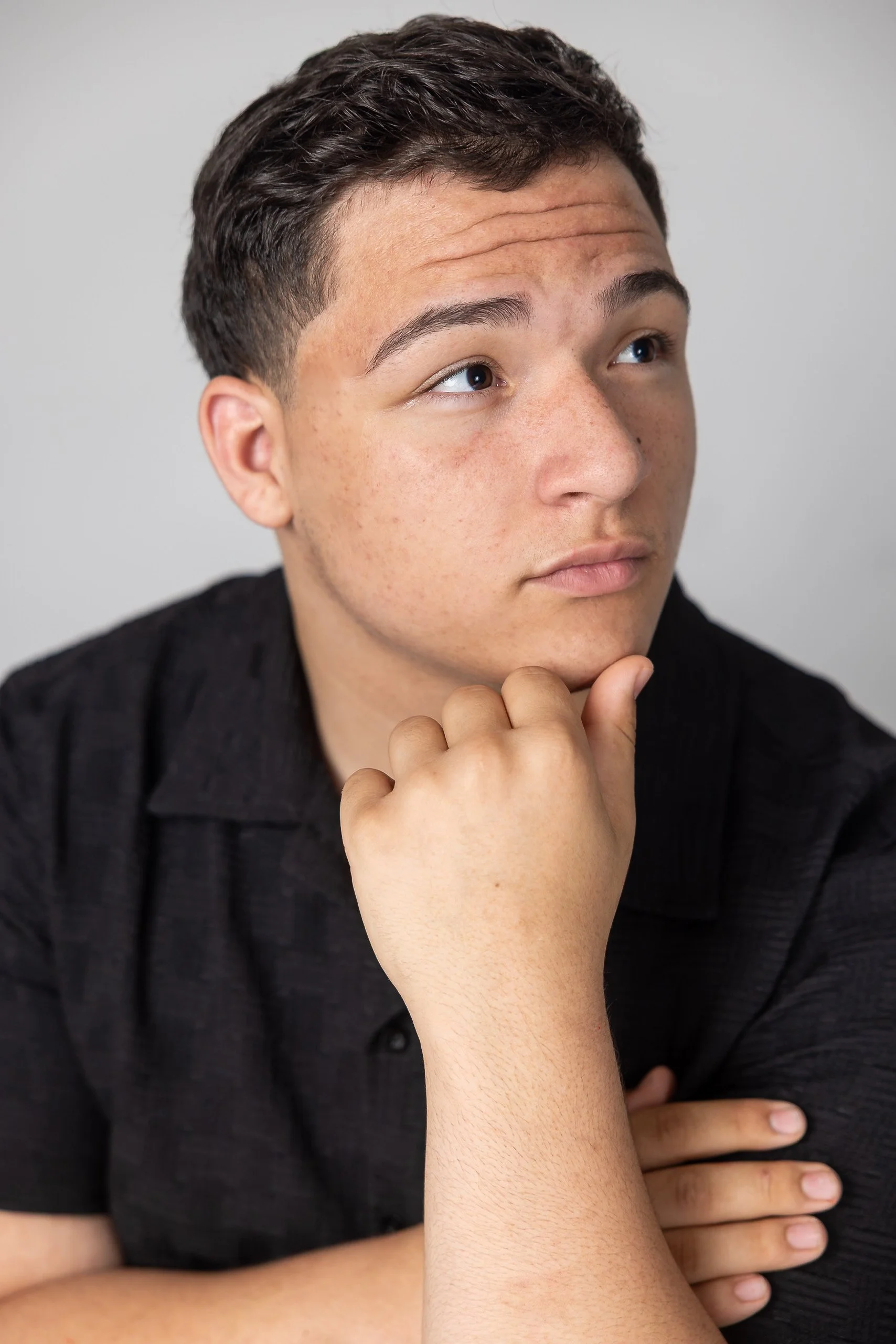 Close-up of a young man with short dark hair, sitting and resting his chin on his hand, wearing a black shirt, against a plain light background.