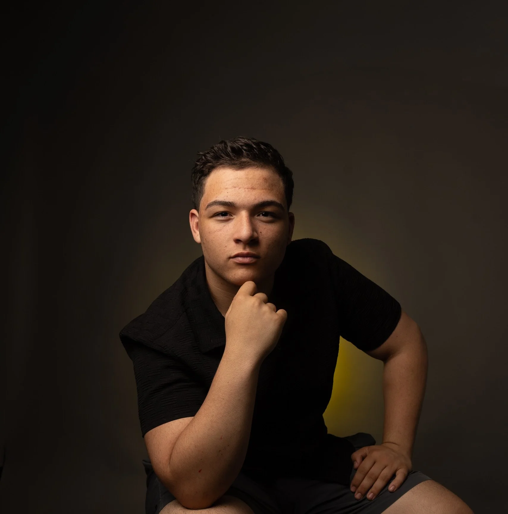 A young man with short dark hair and tattoos on his arm, wearing a black shirt and gray shorts, is posing against a dark background with a yellow gradient. He is resting his chin on his hand, looking directly at the camera with a serious expression.
