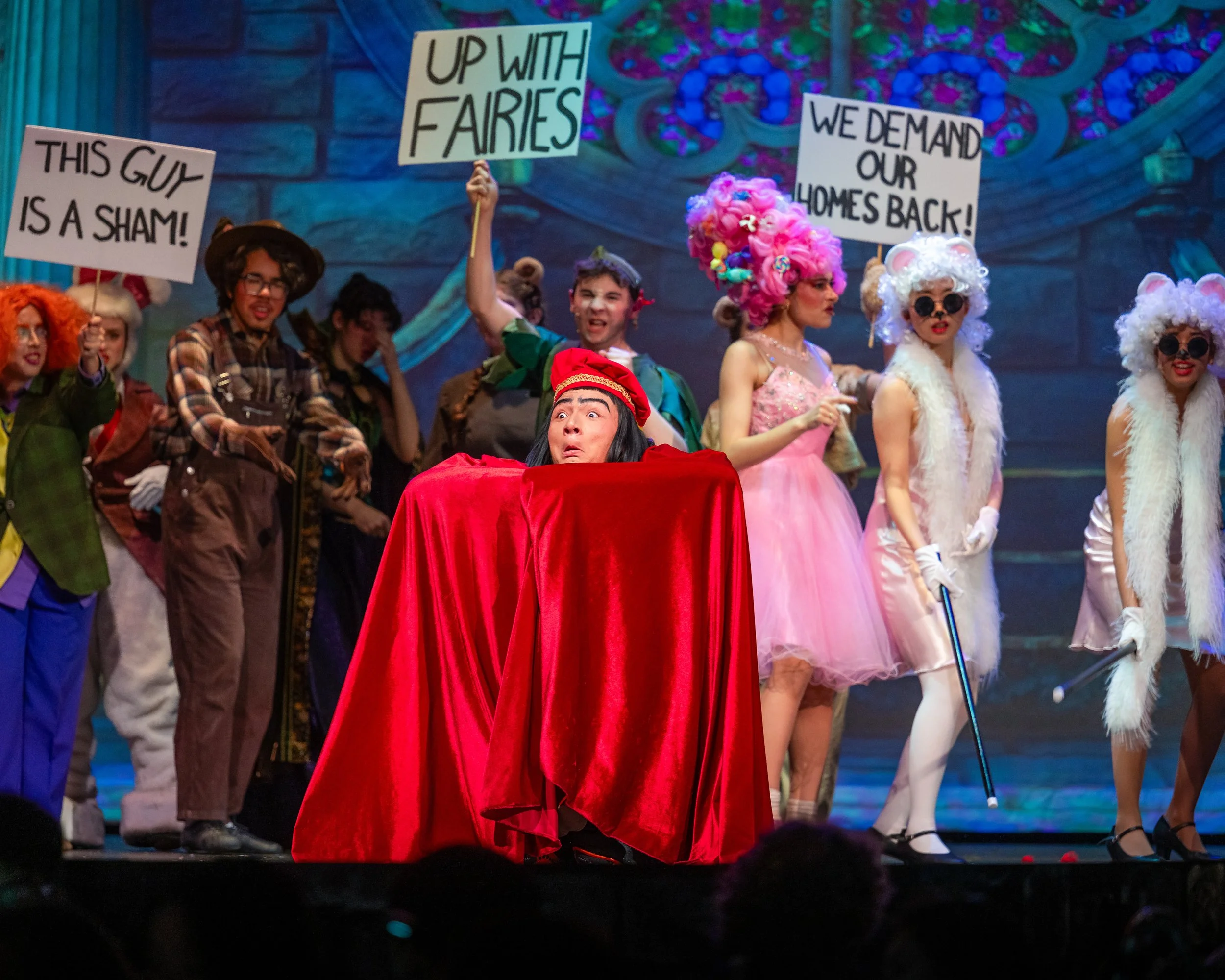People dressed in colorful costumes with signs protesting for fairy rights, in a theatrical setting.