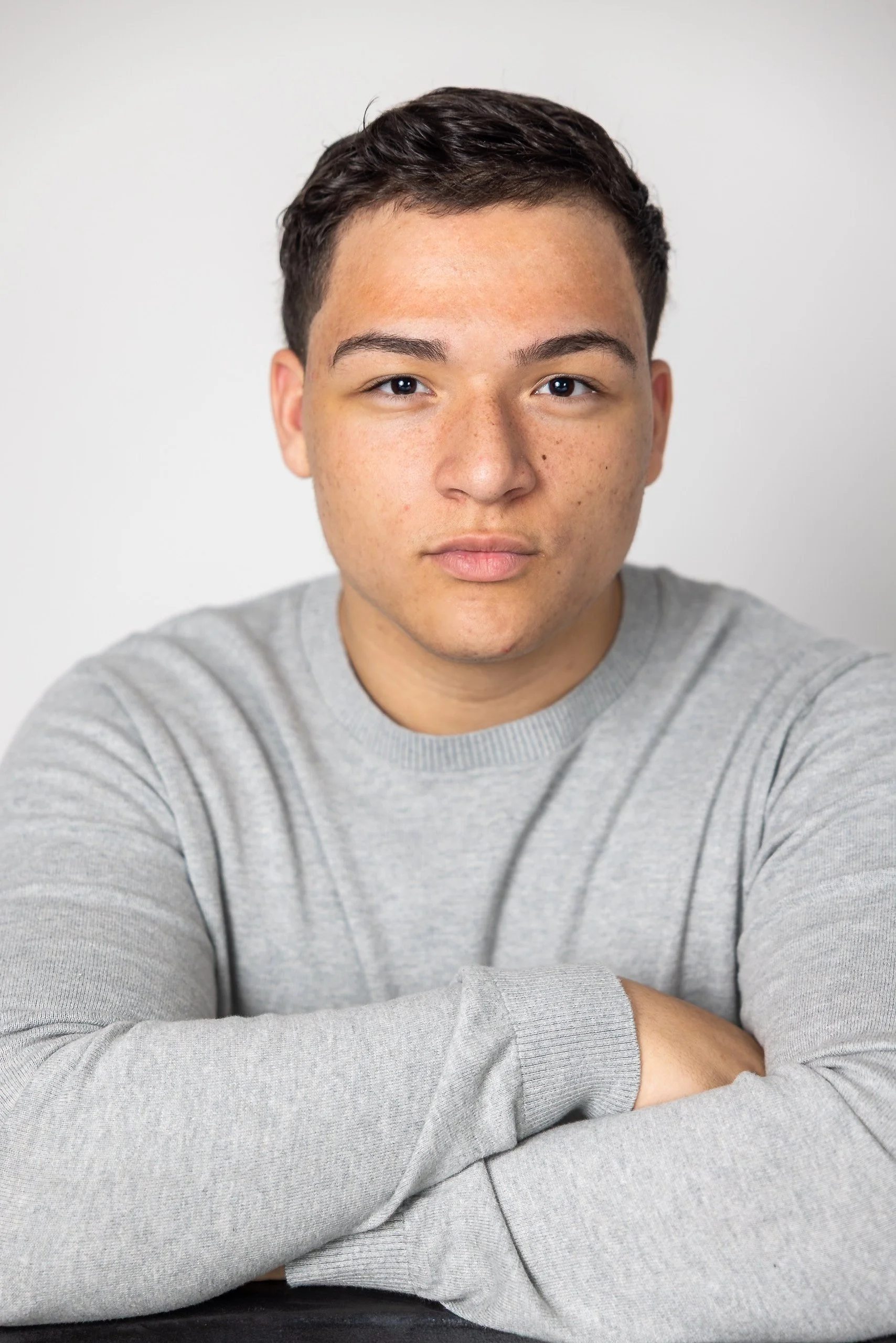 A young man with short dark hair and light skin sitting with arms crossed, wearing a light gray sweater against a plain white background.