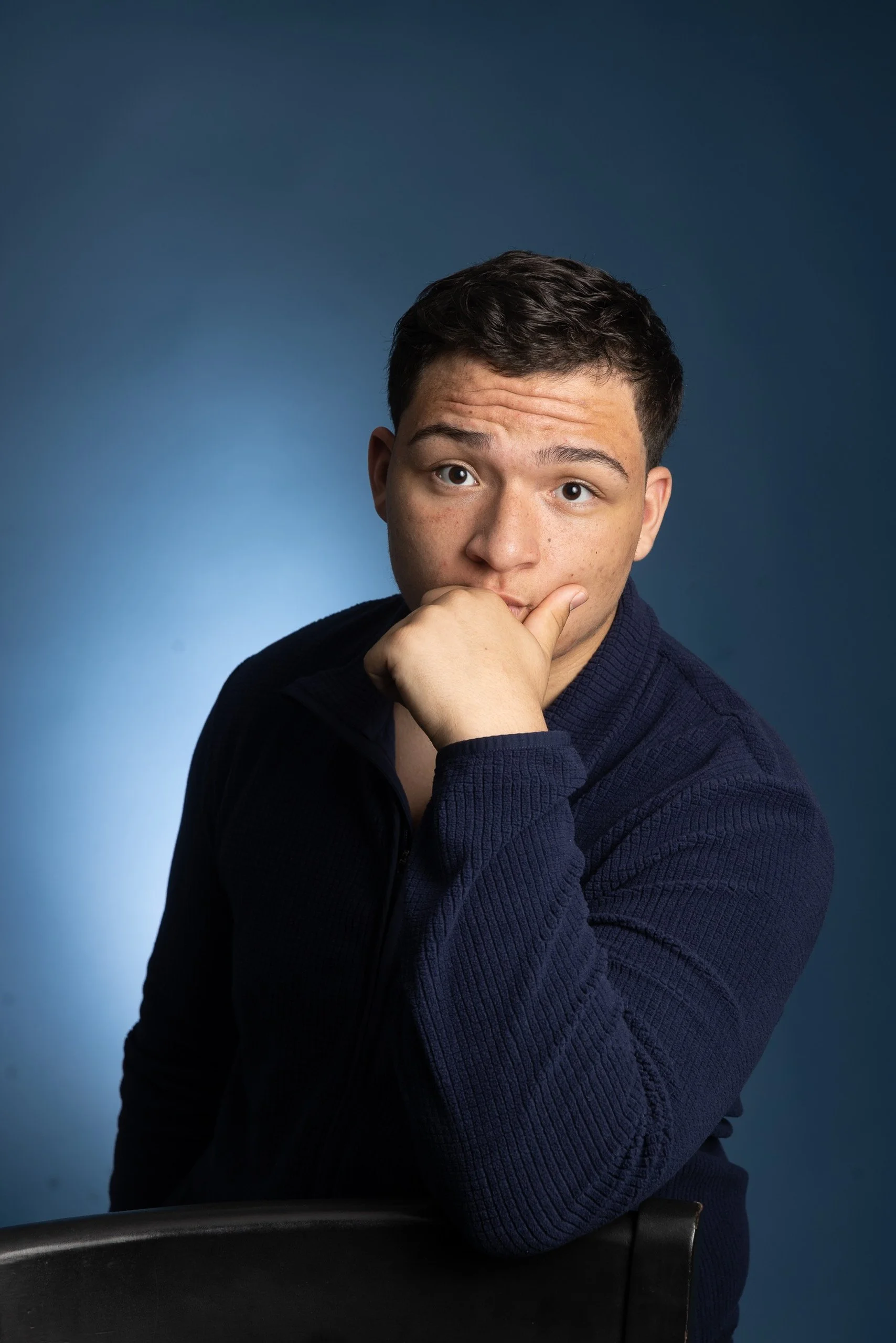 A young man with short dark hair and light skin, wearing a navy blue knitted sweater, is sitting with his hand resting on his chin, looking directly at the camera against a blue background.
