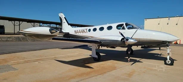 A small private jet airplane parked on tarmac with hangar and blue sky in background.