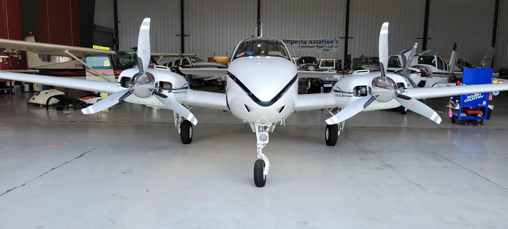 Front view of a small aircraft inside a hangar, with other aircraft and aviation equipment in the background.