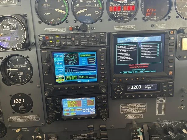 Inside an aircraft cockpit, showing multiple flight instruments, GPS and navigation screens, engine gauges, and control switches.