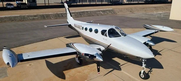 White private propeller aircraft parked on tarmac with a runway and fence in the background.