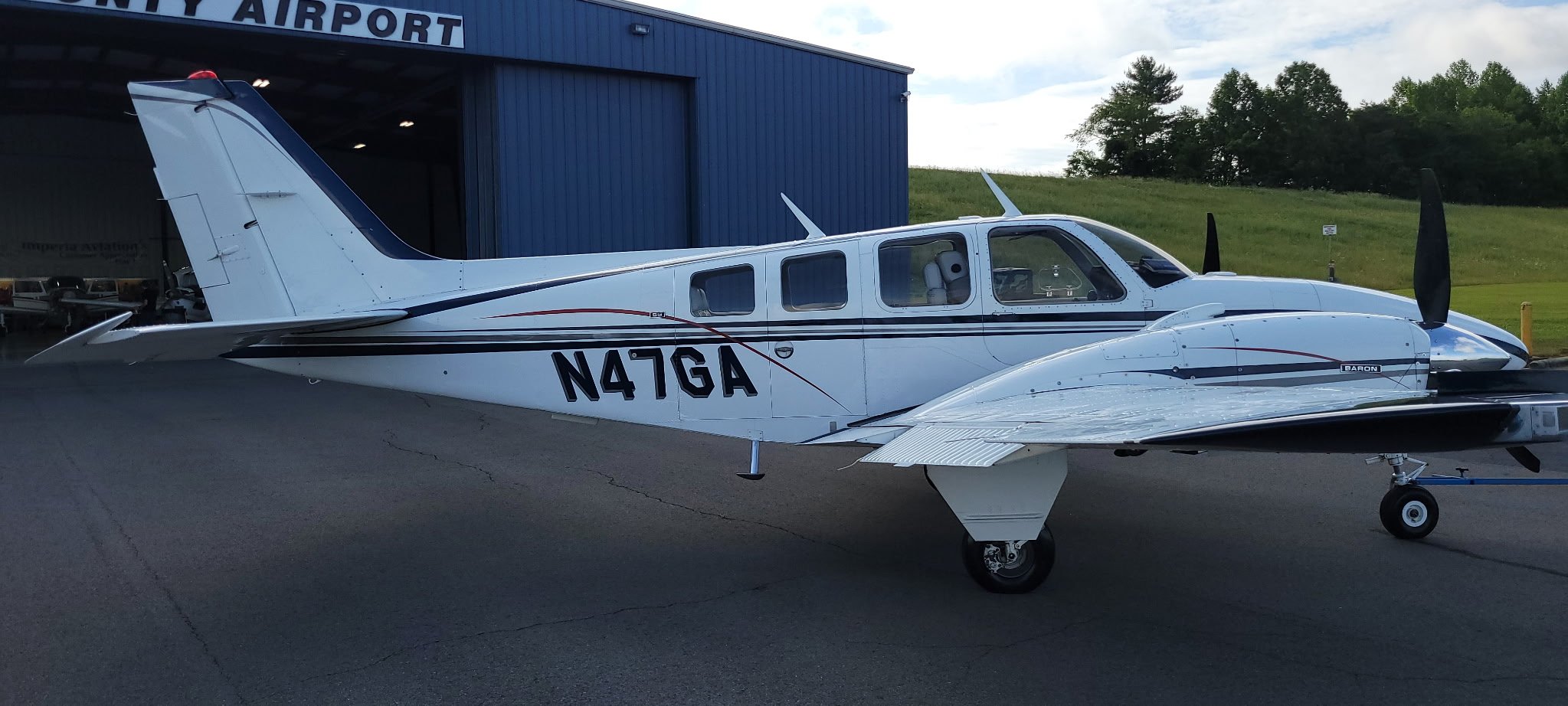 A white private jet with tail number N47GA parked outside a blue aircraft hangar with the name "Airpor" visible at the top. The aircraft has black and red stripes along the side and is positioned on a paved surface with a grassy hill and trees in the background.