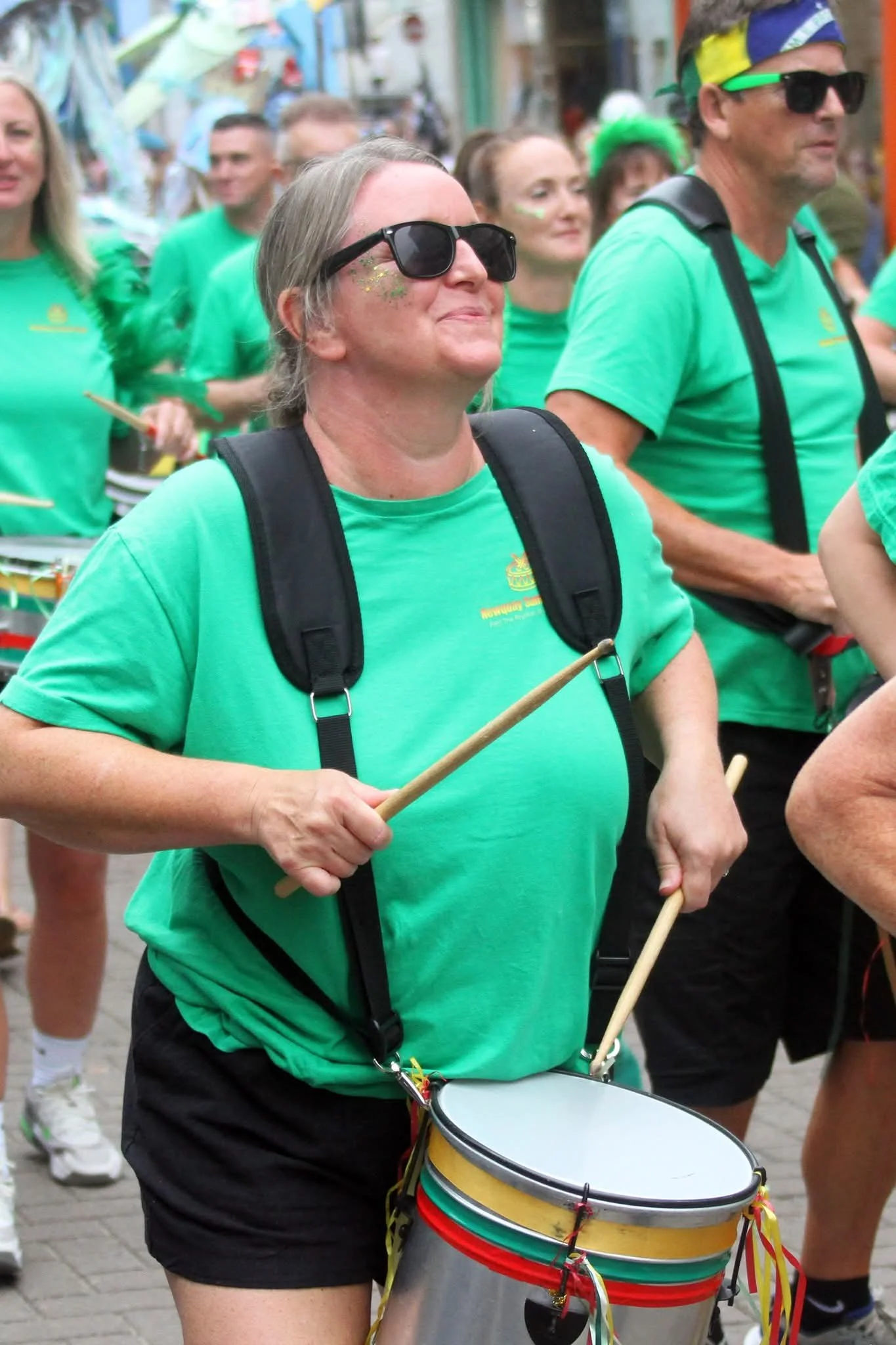 A woman with gray hair, wearing sunglasses, a green T-shirt, and black shorts, playing a marching snare drum with colorful ribbons, participating in a parade or festival with other people wearing green shirts.