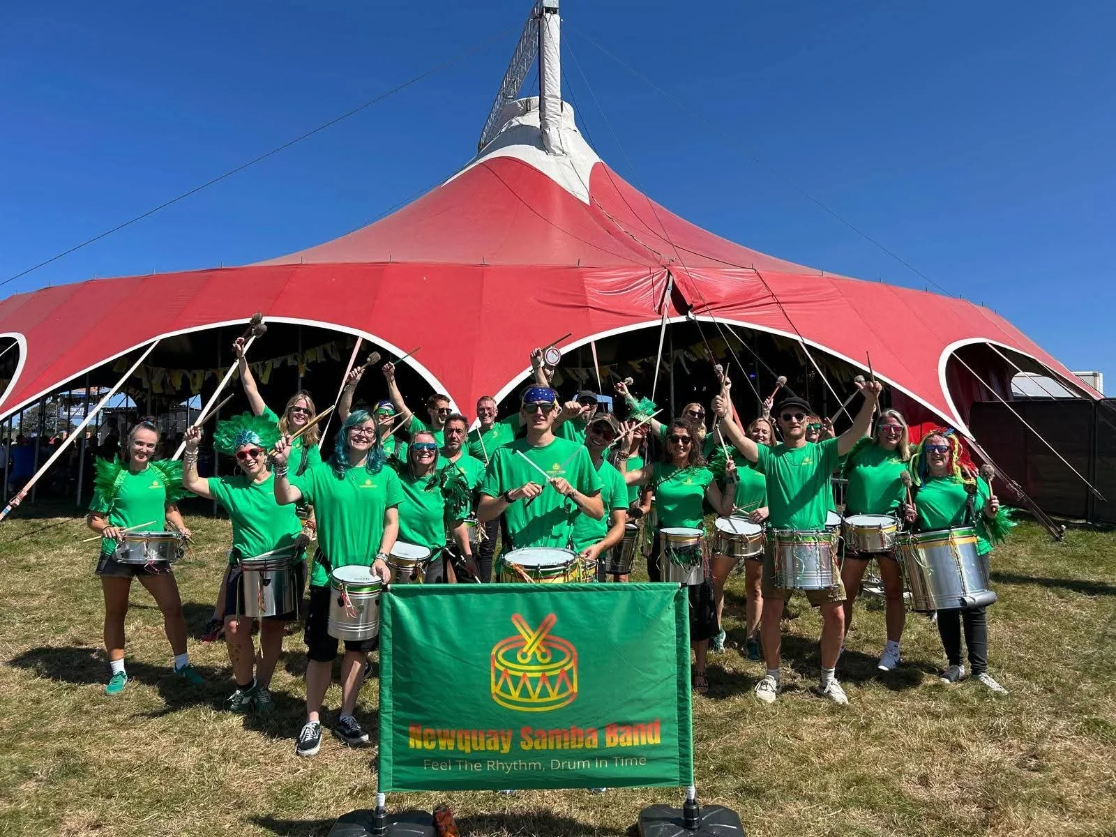 A group of people wearing green shirts and holding drums, standing in front of a large red tent at an outdoor event, with a banner reading 'Newquay Samba band' and a subtitle 'Feel The Rhythm, Drum In Time'.