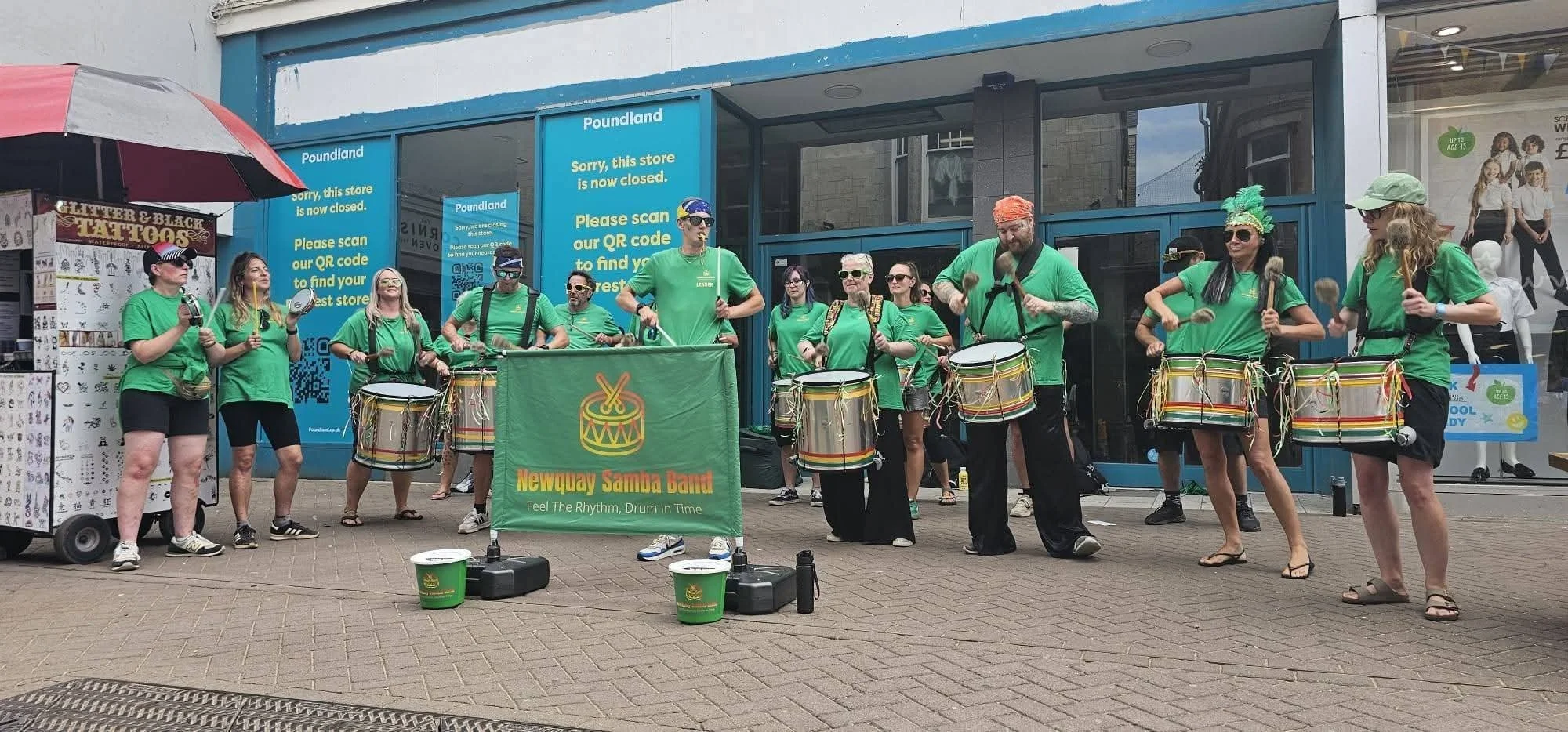 A group of people in green shirts performing as the Newquay Samba Band on a sidewalk outside a closed store, with some holding drums and percussion instruments.