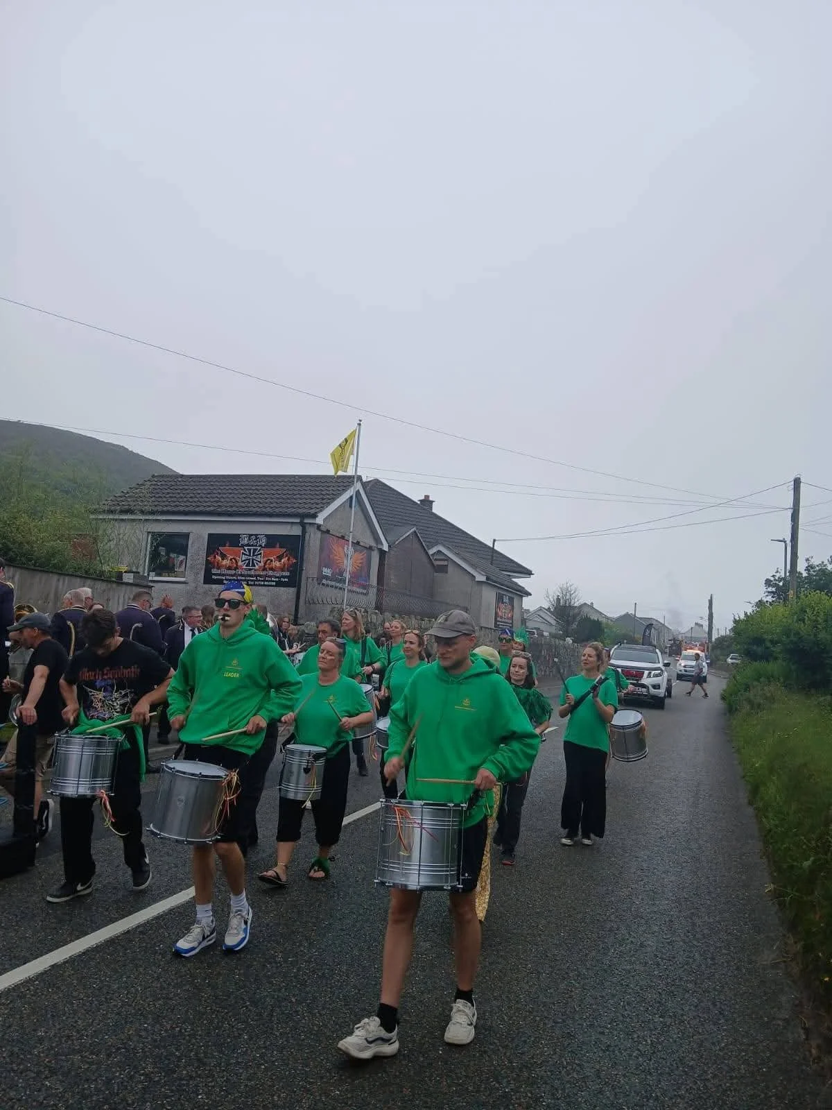 People marching in a parade with drums, wearing green hoodies, on a street in a residential area with houses, cars, and utility poles visible.