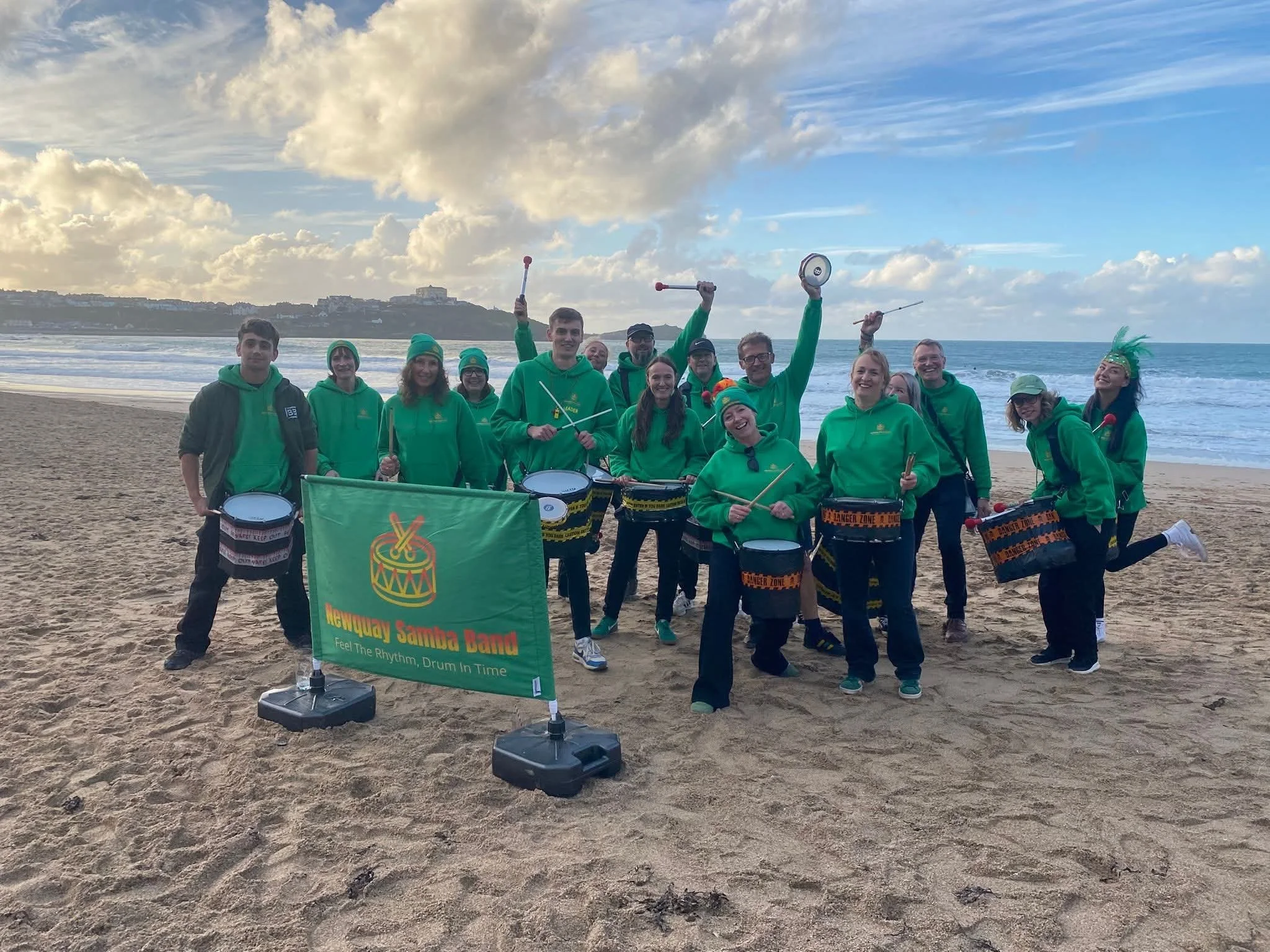 A group of people wearing matching green hoodies and hats, holding musical instruments, on a sandy beach with ocean waves, cloudy sky, and distant land in the background.