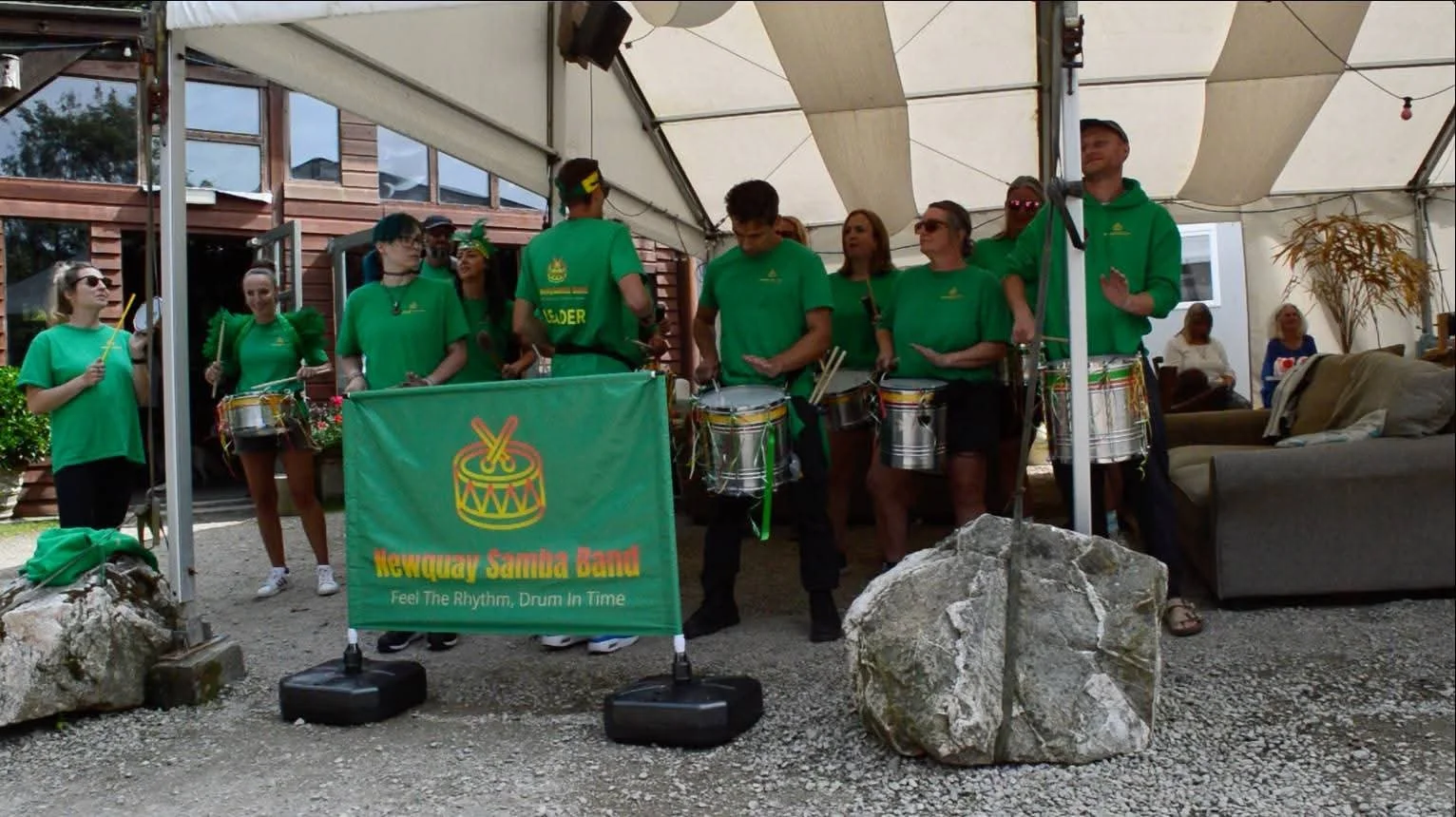 A group of people wearing green shirts, playing drums under a tent at an outdoor event. A banner in front reads 'Hawhey Samoa Band,' with the tagline 'Feel The Rhythm, Drum In Time.'