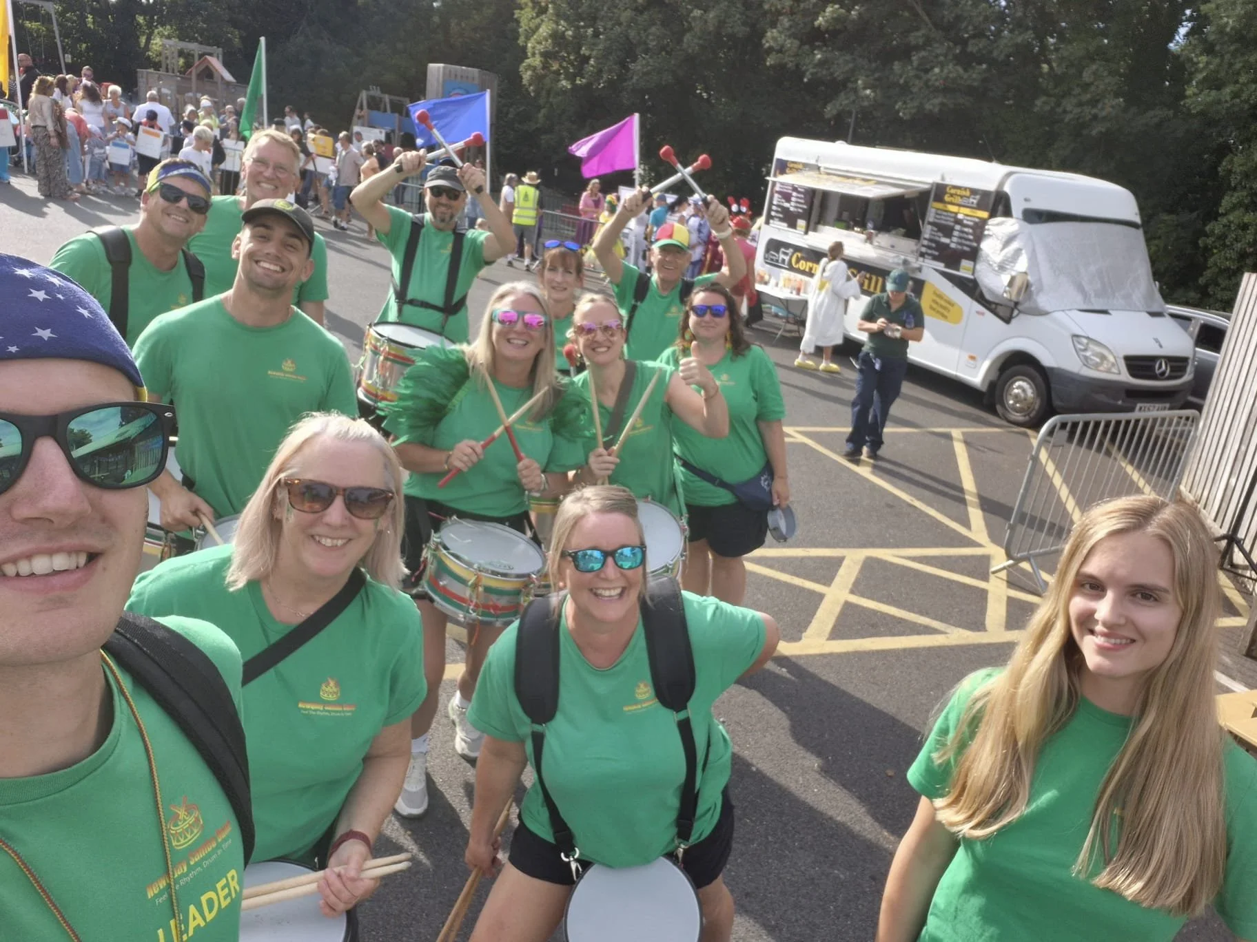 Group of people wearing green shirts participating in an outdoor festival or parade, some holding drums and drumsticks, with food trucks and other attendees in the background.