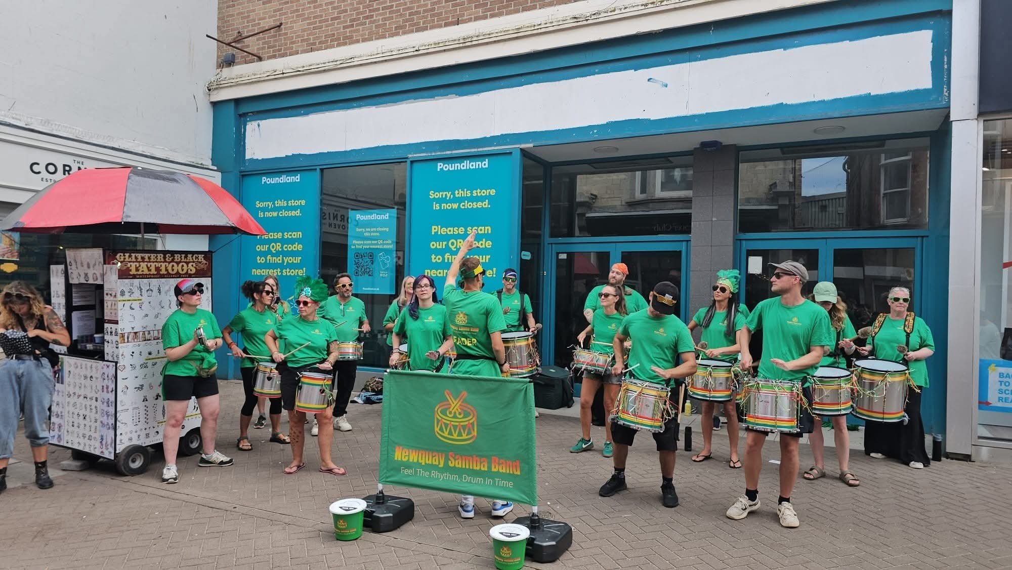 Street scene outside a closed store with a group of people in green shirts playing drums for a band called 'Newquay Samba Band.' The store has yellow signs indicating it is closed, with scanning instructions for a QR code. A woman with a pink and bla
