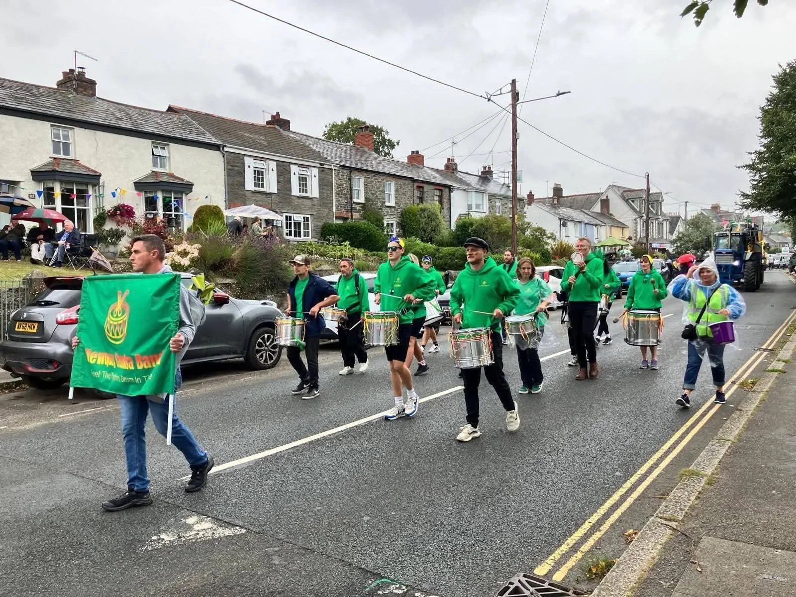 People participating in a parade walking down a street, one person at the front holding a green flag reading 'Cwmdu Band,' with others playing drums and carrying umbrellas on a cloudy day in a residential neighborhood.