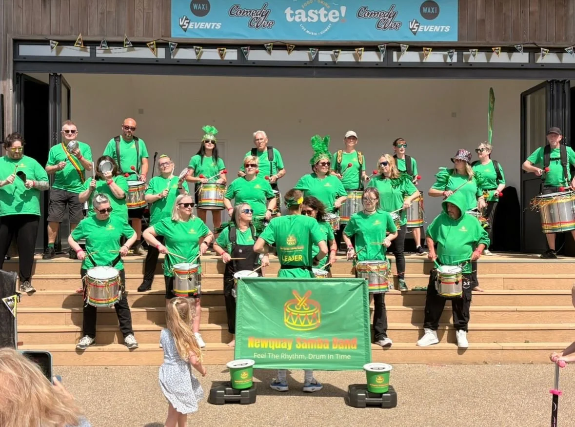Group of people wearing green shirts, playing drums on a stage during a parade or event. There is a banner that reads 'Newquay Samba Band' and a sign above the stage with logos and text.