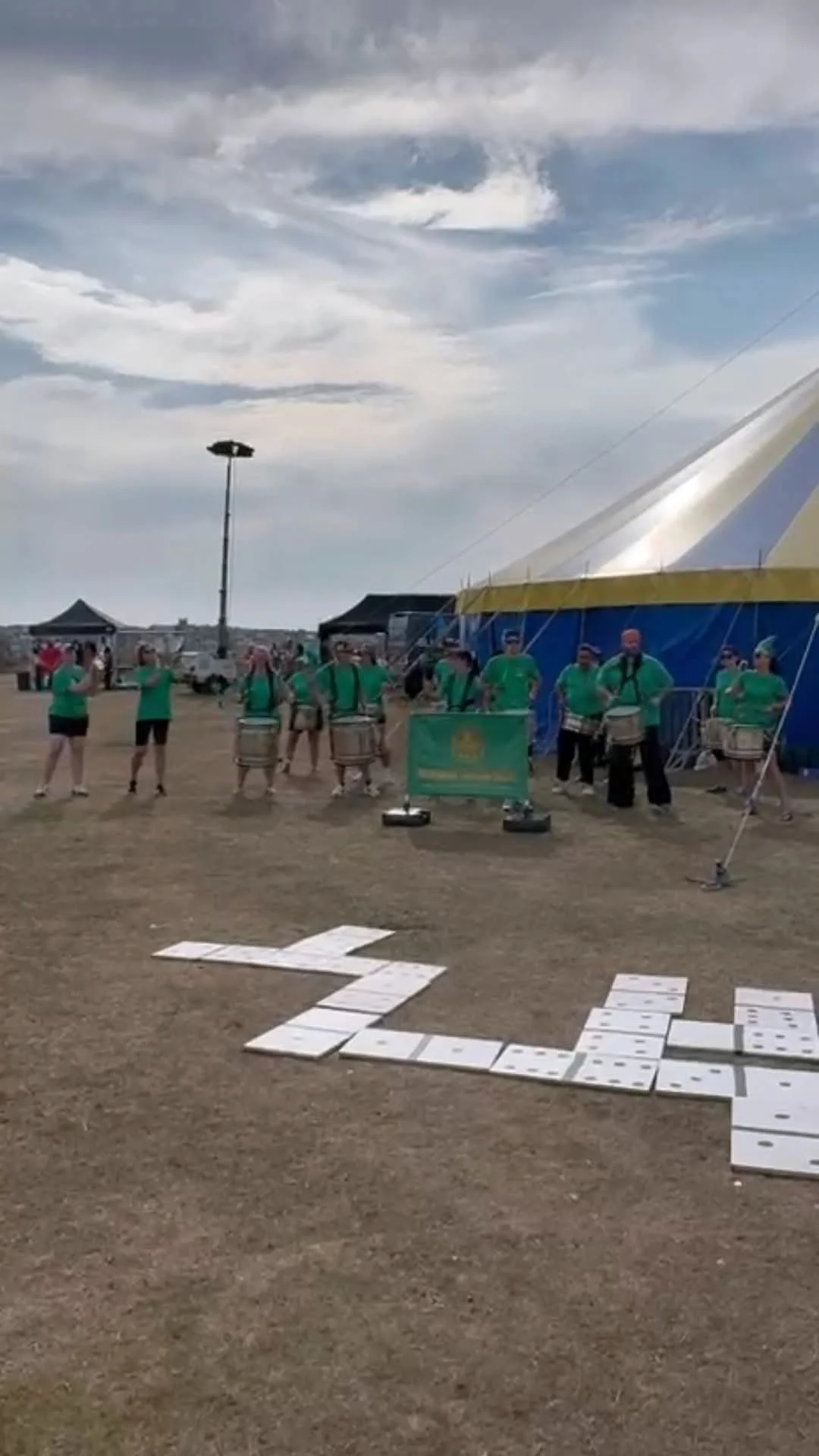 A group of people in green shirts standing near a large blue and yellow circus tent, with some playing drums and others taking photos, under a cloudy sky.
