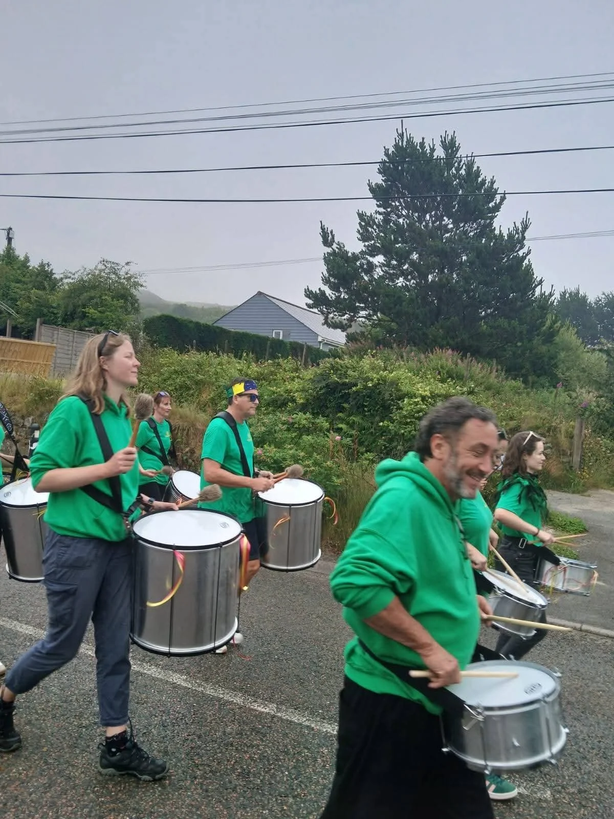 A group of people walking outdoors, dressed in green shirts, playing drums, and smiling, with a background of trees, bushes, a house, and overcast sky.