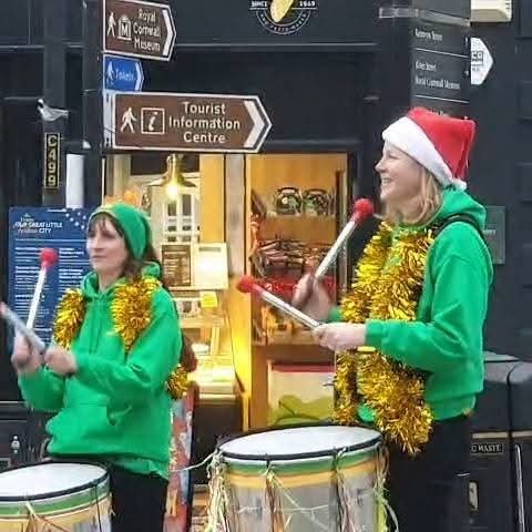 Two women dressed in green hoodies, holiday hats, and gold tinsel scarves playing drums outdoors, with one woman wearing a Santa hat and the other a green hat with a pom-pom.