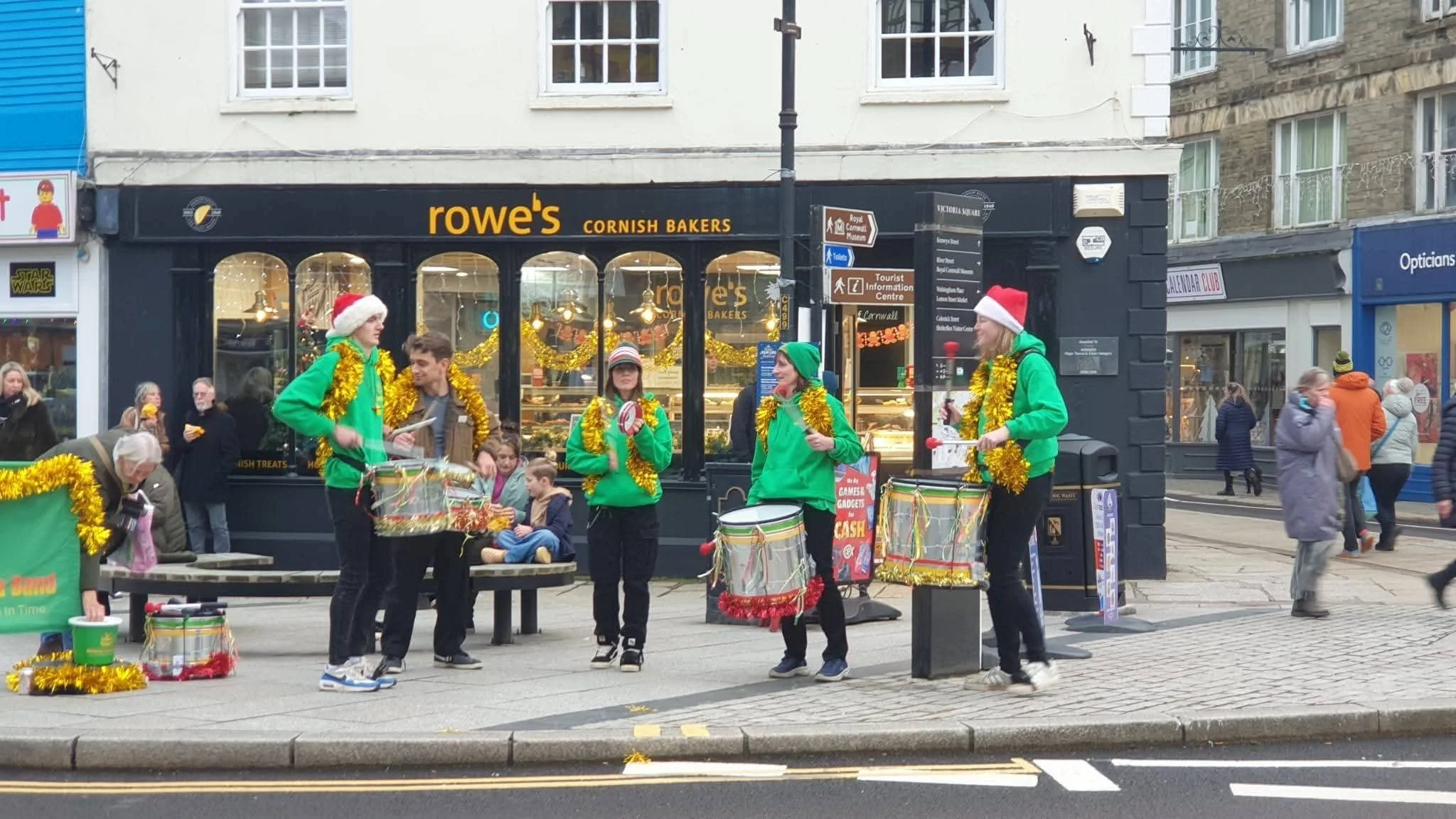 Street scene with a bakery storefront named 'rowe's CORNISH BAKERS' decorated for Christmas. Several people, some wearing Santa hats and green jackets with gold tinsel, are playing drums and engaging with a small crowd. Pedestrians walk by, and build