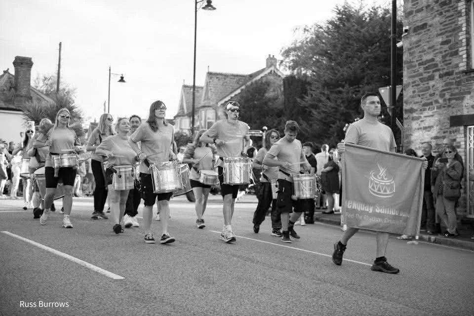 A parade featuring children and adults marching down a street, with some playing drums and one carrying a flag. Spectators are watching from the sidewalk.