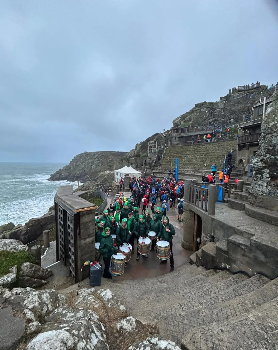 A large group of people gathered on a coastal outdoor venue with stairs, some holding umbrellas, near the ocean under a cloudy sky. There is a marching band with drums, and the setting appears to be at a seaside with rocky cliffs.