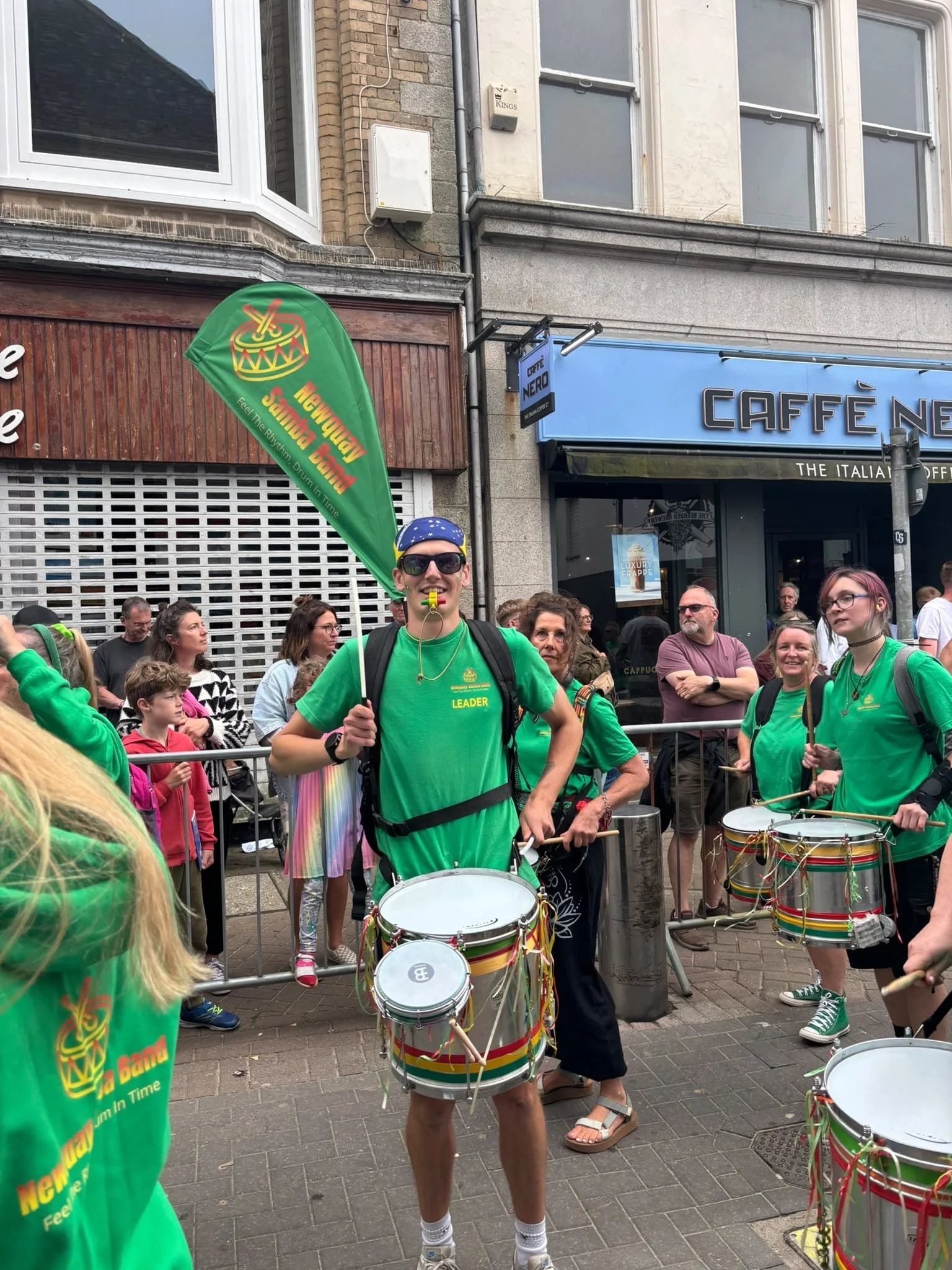 A group of people participating in a street parade or festival, some playing drums and others watching, with buildings and shop signs in the background.