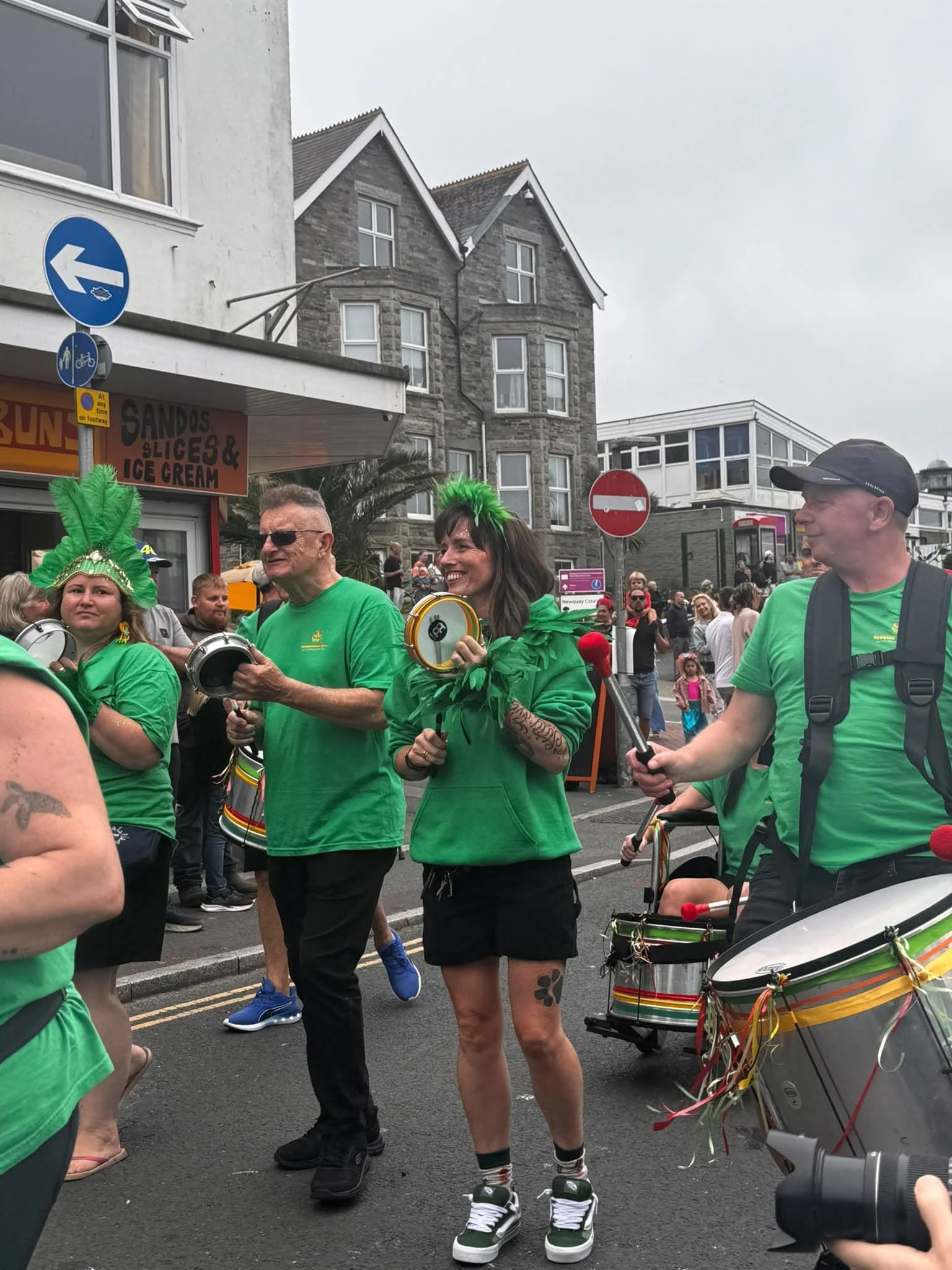 People participating in a parade or street celebration, wearing green clothing, some with feathered headpieces, and playing drums. The scene takes place on a city street with buildings and onlookers in the background.
