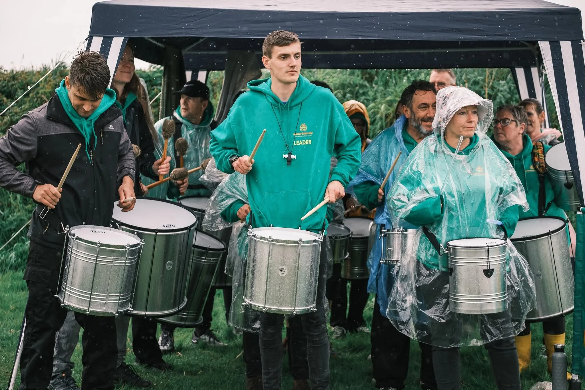 Group of people playing drums outdoors under a canopy on a rainy day.