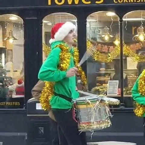 Person wearing a Santa hat and green jacket, carrying a drum and holiday decorations, in front of a shop window with Christmas ornaments.