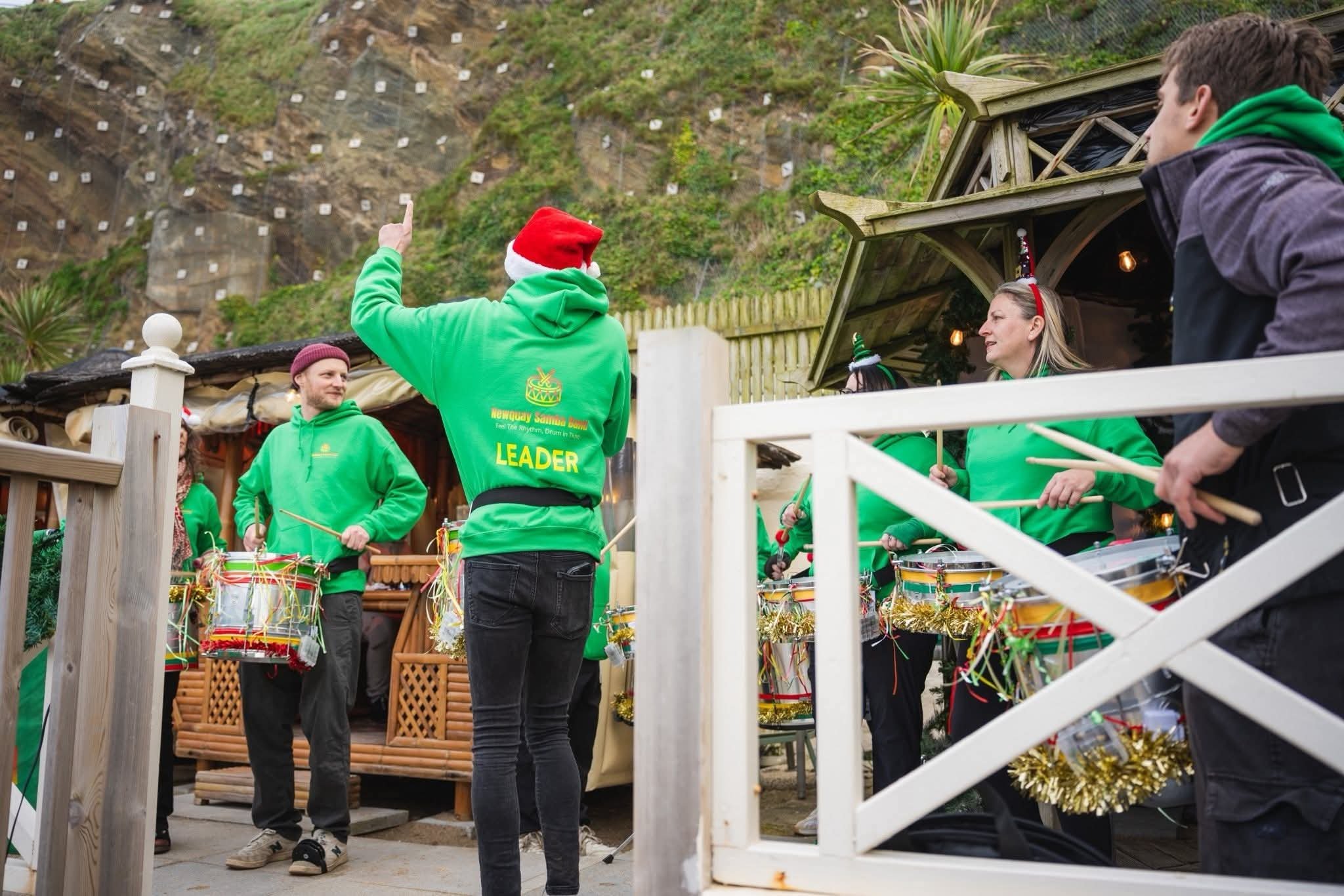 A group of performers wearing green hoodies, some with Santa hats, preparing for a Christmas parade with drums decorated with gold tinsel, as a hillside with trees and houses is visible in the background.