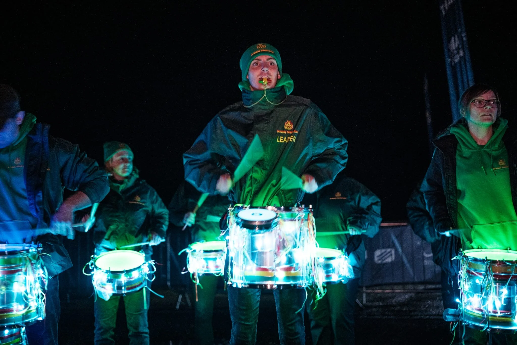 Group of people playing drums at night, illuminated by colorful lights, with a dark sky background.