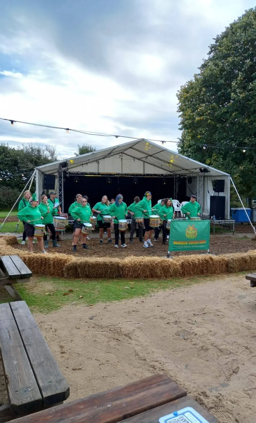 A group of people wearing green shirts playing drums on an outdoor stage at a festival or event, surrounded by hay bales and trees under a partly cloudy sky.