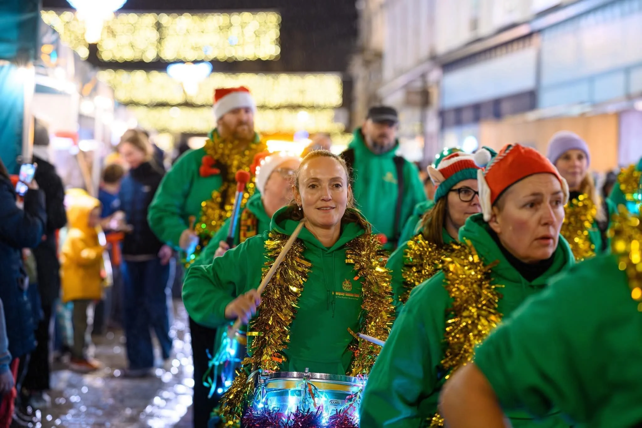 People dressed in green and wearing Santa hats, participating in a festive parade with tinsel and lights, on a city street at night.